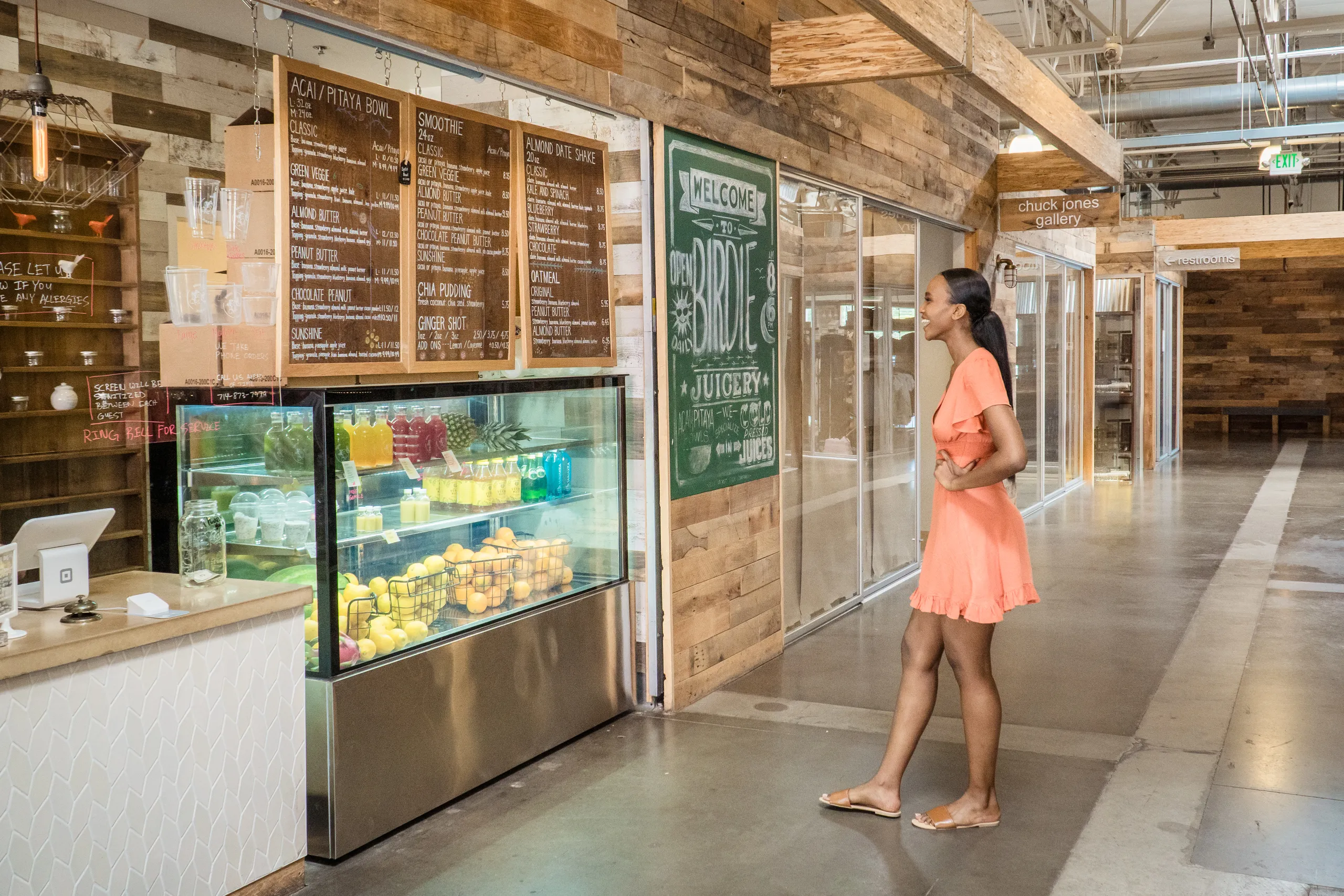 A woman in an orange dress stands in front of a juice bar counter, looking at the menu. The shop displays fresh fruit and bottled juices in a glass case. The setting is modern with wood accents.