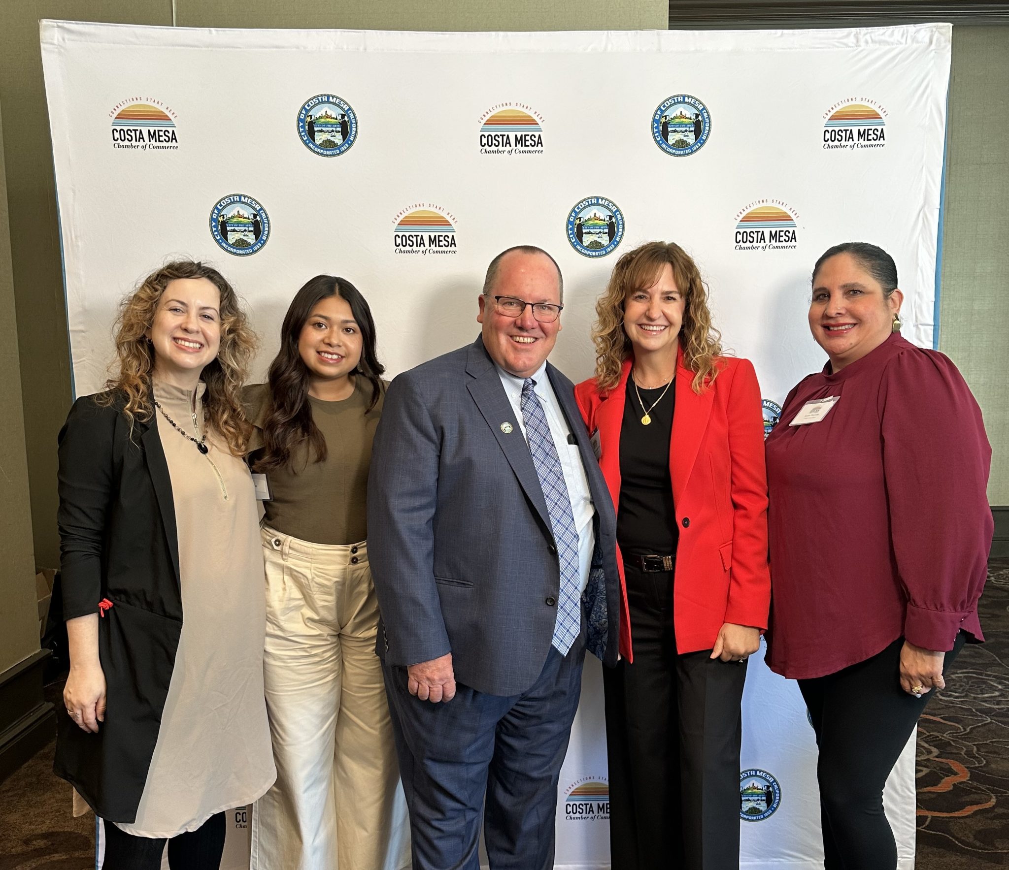 Five people stand smiling together in front of a white backdrop with “Costa Mesa” and city seal logos, dressed in professional attire at an event.
