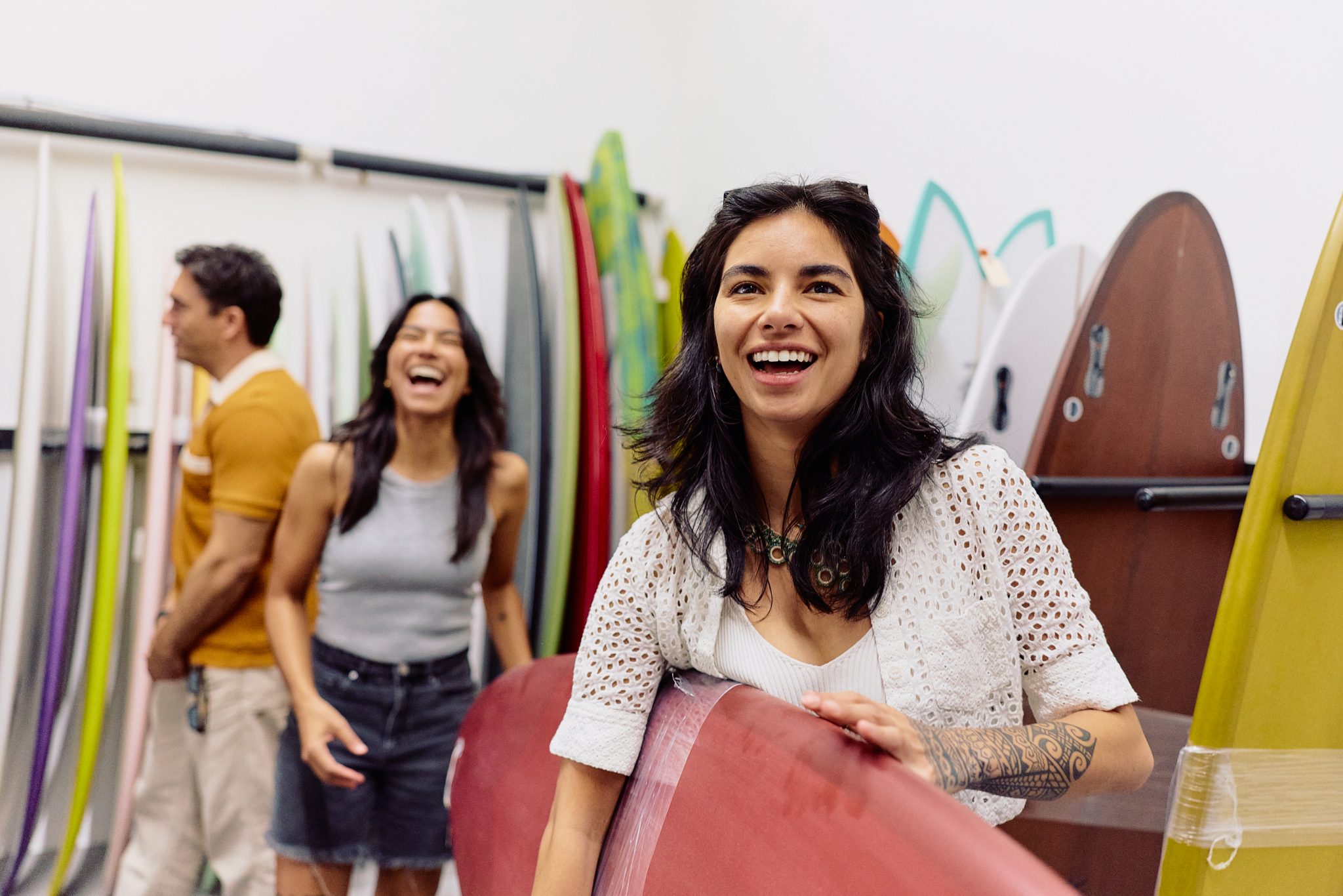 Three people smile and laugh while standing among colorful surfboards in a shop. A woman in the foreground holds a red surfboard, with two others happily chatting in the background.