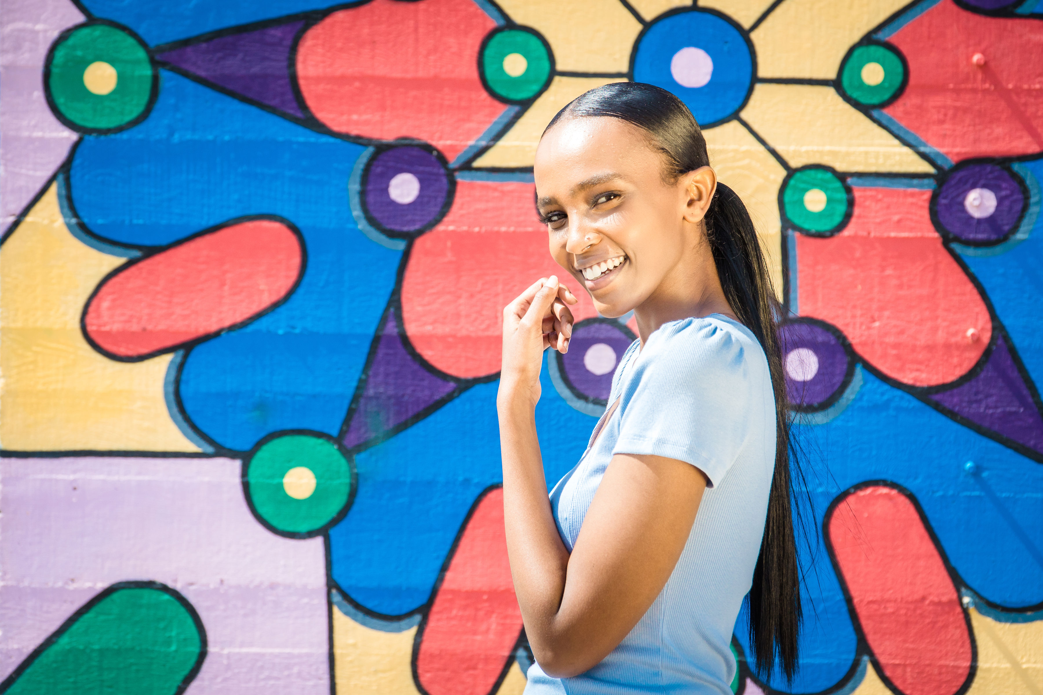 A smiling woman in a light blue shirt stands before a colorful, abstract mural in Costa Mesa, City of the Arts, with geometric shapes and bright hues, looking at the camera with her hand near her face.