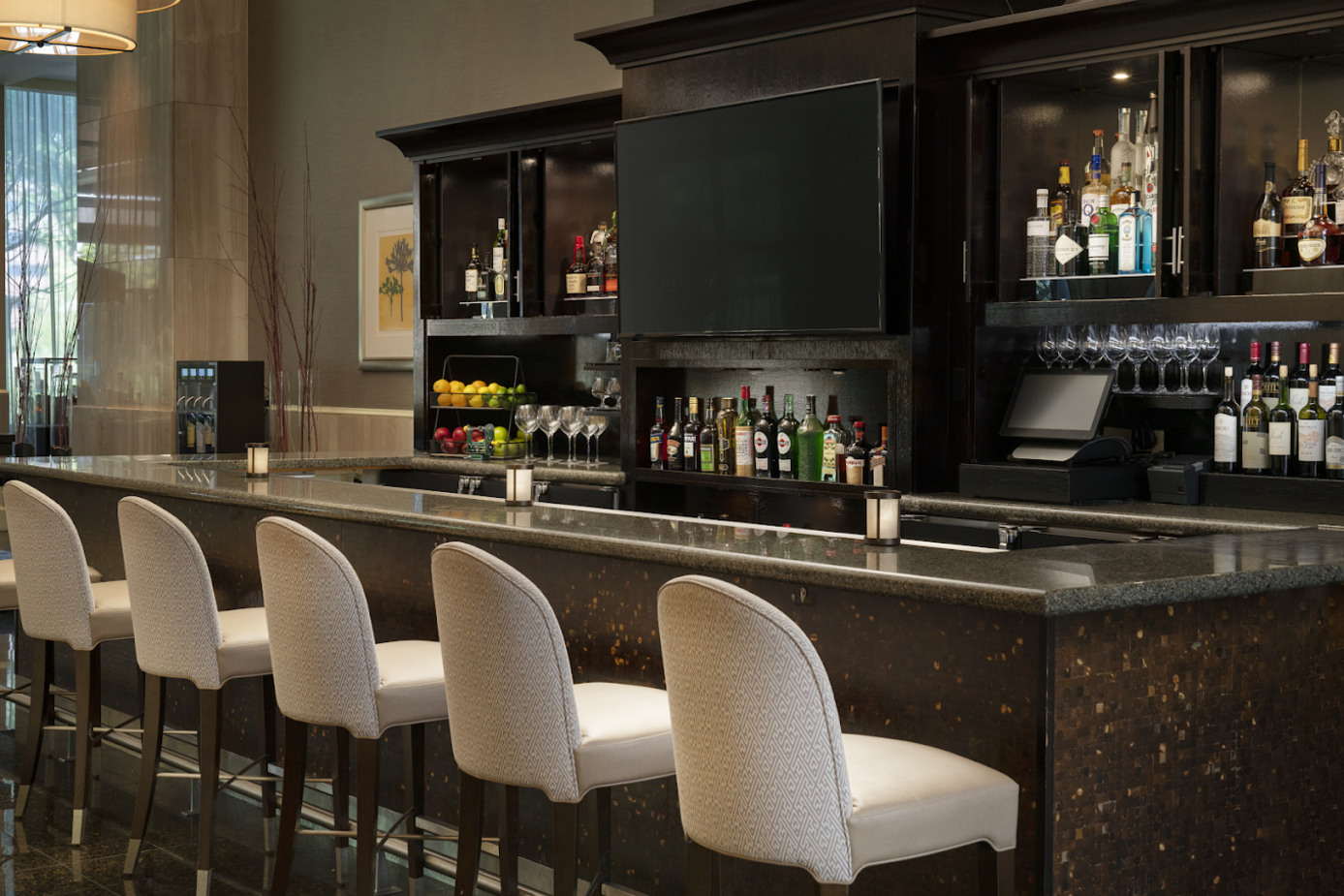 A modern hotel bar with six white upholstered stools, a dark countertop, shelves of liquor bottles, wine glasses, and a large TV screen above the counter. The area is well-lit and stylishly decorated.