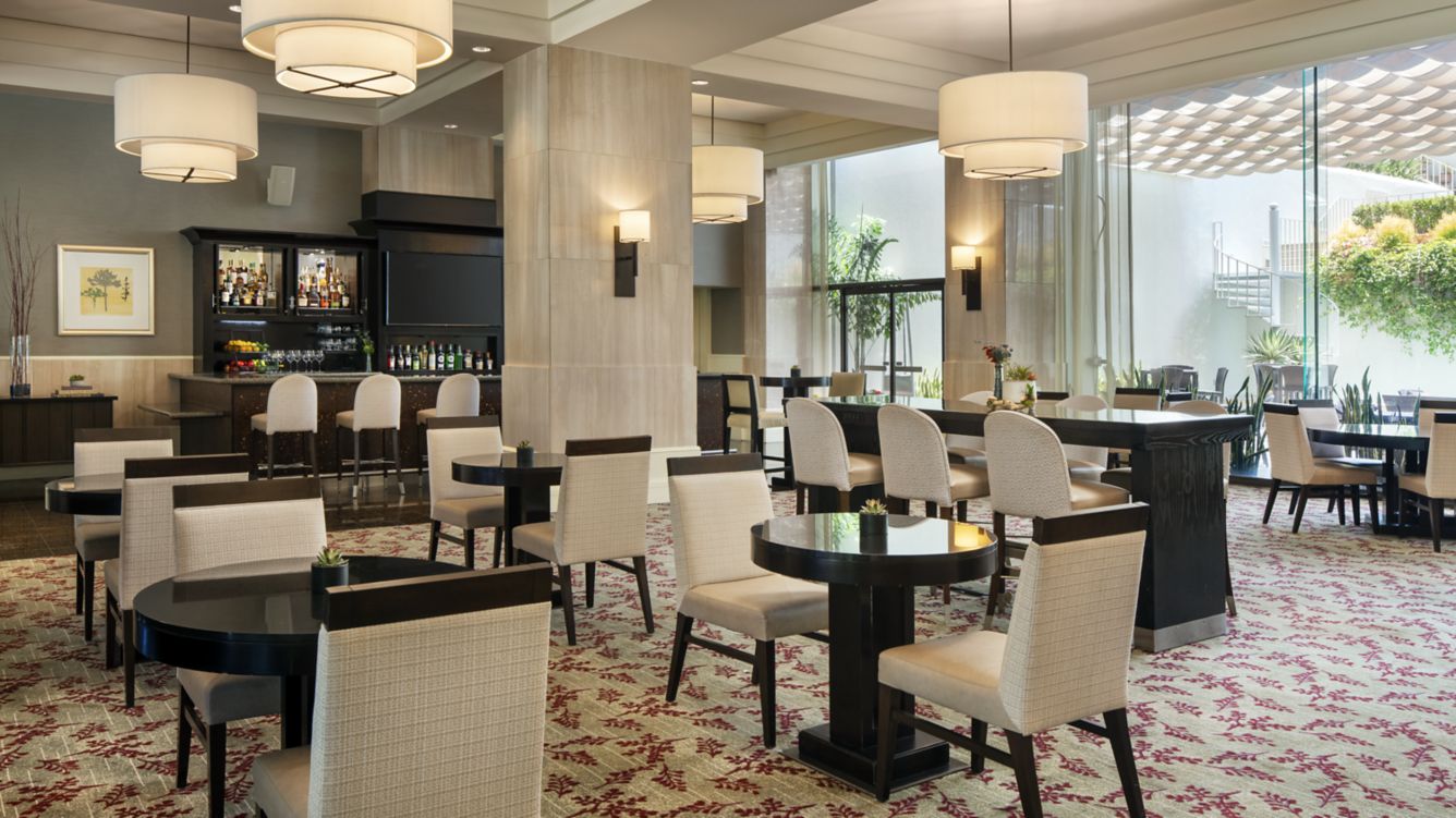 A modern, spacious restaurant with round tables, cushioned chairs, large hanging lights, and a bar in the background; natural light streams in through tall windows with greenery visible outside.