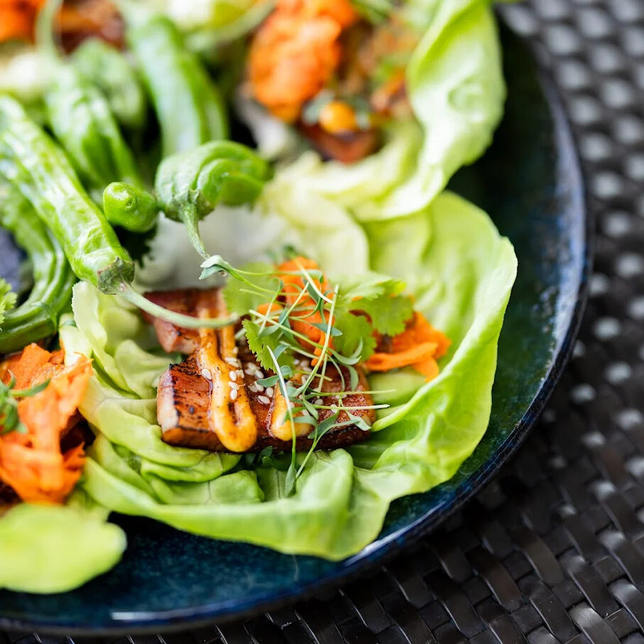 A close-up of lettuce wraps filled with grilled tofu, shredded carrots, fresh cilantro, greens, and sauce, arranged on a dark plate.