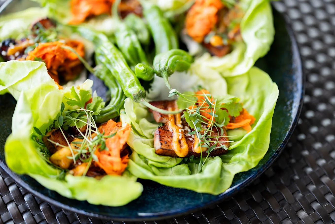 A close-up of lettuce wraps filled with grilled tofu, shredded carrots, fresh cilantro, greens, and sauce, arranged on a dark plate.