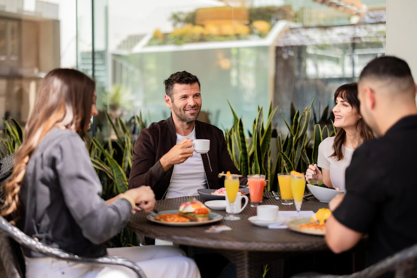 Four people sit around a table outdoors, enjoying breakfast with plates of food, coffee cups, and juice glasses. They are smiling and talking, surrounded by greenery and modern buildings in the background.