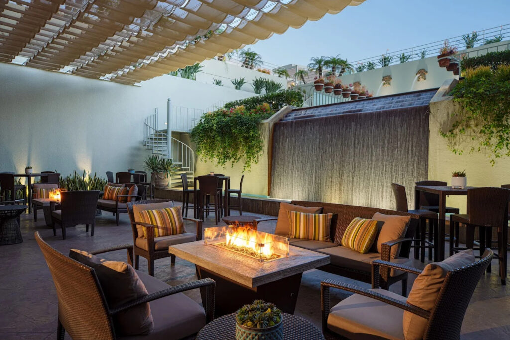 Modern outdoor patio with cushioned seating around a fire pit, tall tables, potted plants, a cascading waterfall feature, a spiral staircase, and a partially covered roof. The space is cozy and inviting.