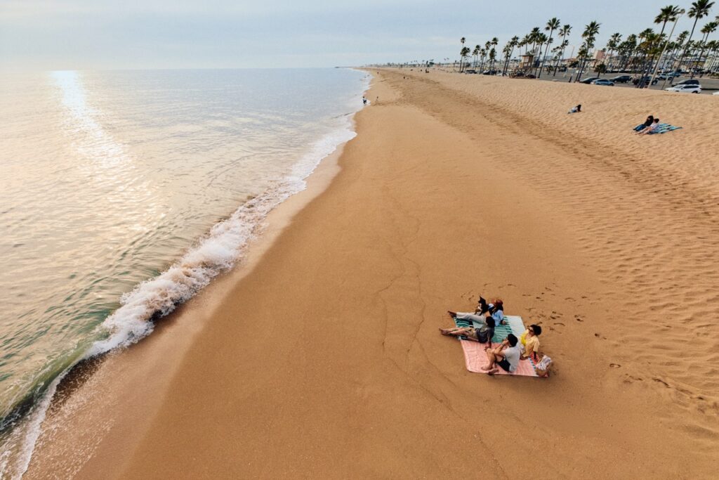 Three people relax on a sandy beach near the waterline, with gentle waves washing ashore and palm trees lining the background. A few other people are spaced along the wide, empty beach under a cloudy sky.