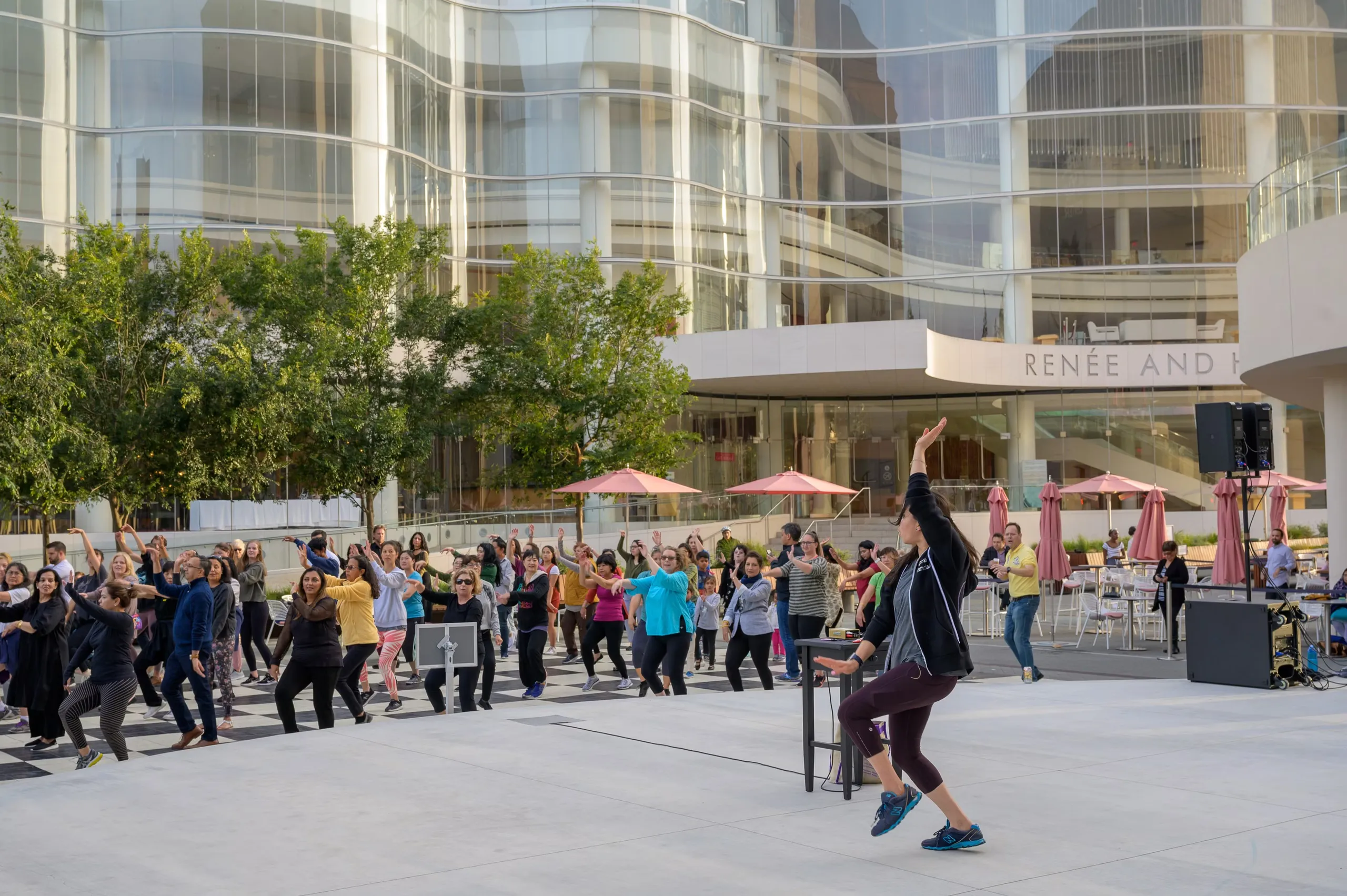 A large group of people participate in an outdoor dance or fitness class led by an instructor, with modern glass buildings, trees, and patio tables with pink umbrellas in the background.