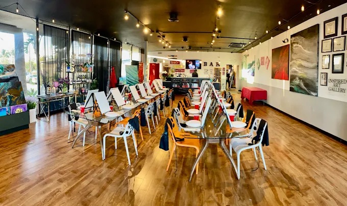 A spacious art studio with wooden floors, easels, and canvases on long tables, surrounded by assorted chairs. Artworks and supplies fill the room, with decorative lights overhead—an inspiring spot among the best things to do in Costa Mesa.