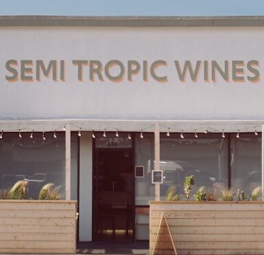 The front of a building with large letters reading SEMI TROPIC WINES. The exterior features light wood planters with greenery and a shaded entrance—perfect for those seeking unique things to do in Costa Mesa.