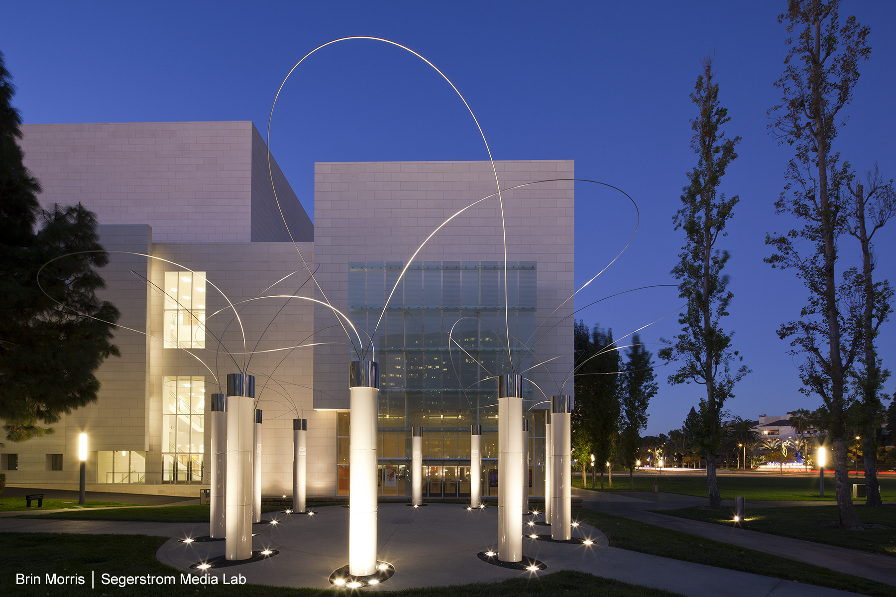 Modern art installation with tall white pillars and curved metal rods stands illuminated in front of a contemporary building with large glass windows, surrounded by trees at dusk—a striking highlight of the Costa Mesa Art Walk.