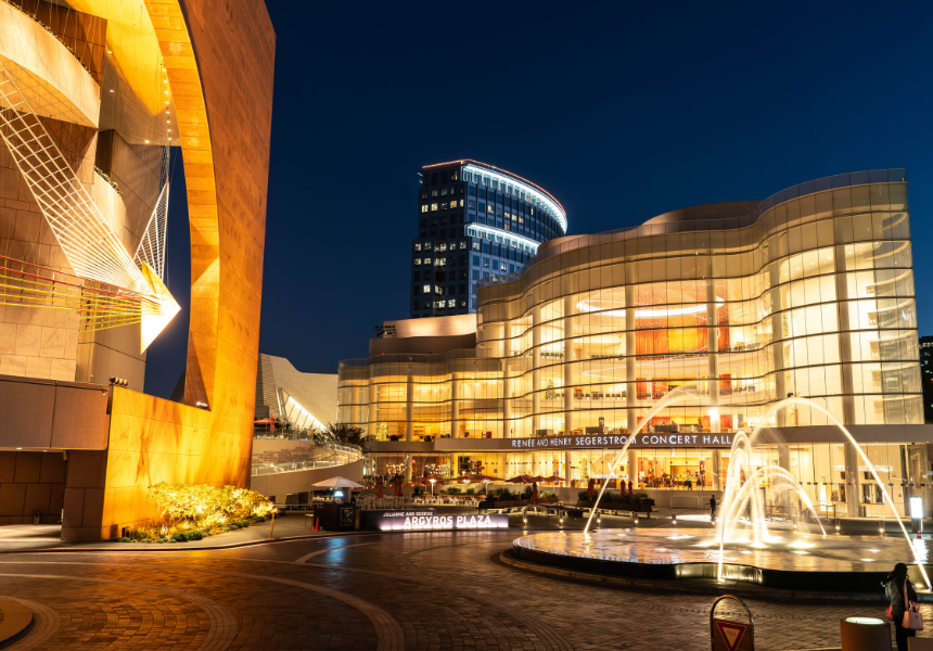 Night view of a modern concert hall with illuminated glass walls, a water fountain in the foreground, and a tall building in the background—capturing the vibrant energy of Los Angeles, set to host FIFA World Cup 2026 events.