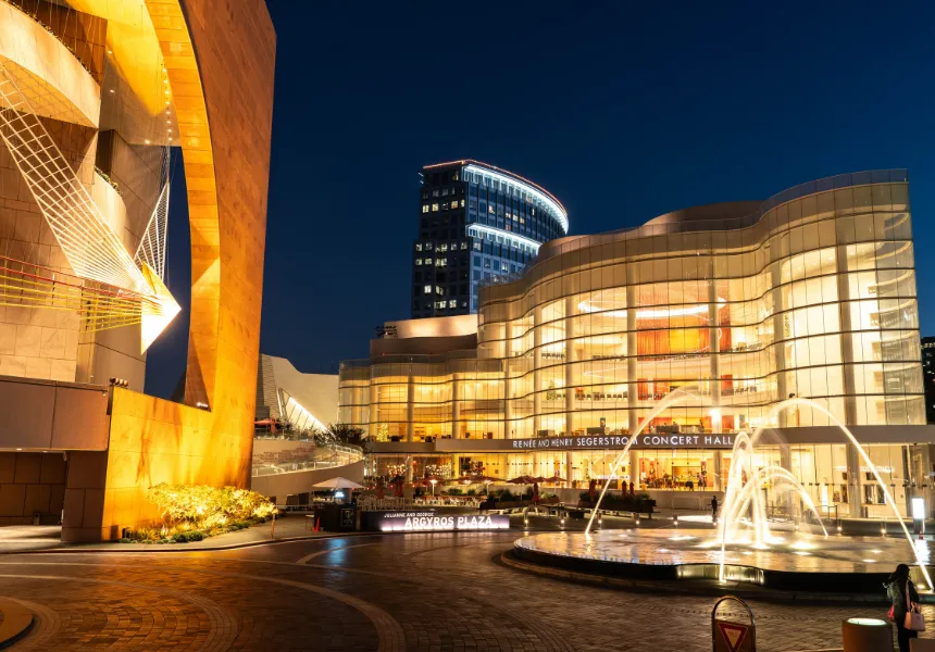 Night view of a modern concert hall with illuminated glass walls, a water fountain in the foreground, and a tall building in the background—capturing the vibrant energy of Los Angeles, set to host FIFA World Cup 2026 events.