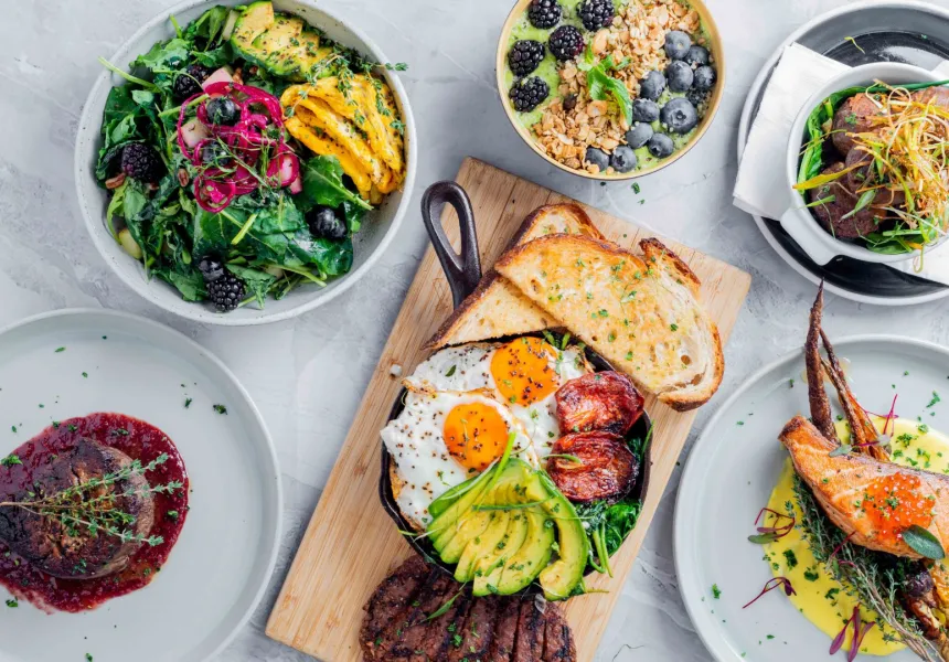 An overhead view of various colorful brunch dishes, including avocado toast with eggs, a salad, a granola bowl with berries, steak with sauce, and salmon, beautifully arranged on a white table—perfect for enjoying during FIFA World Cup 2026 Los Angeles events.