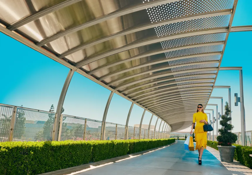 A woman in a yellow dress and sunglasses strolls along a modern covered walkway with shopping bags, under the clear blue sky and manicured green hedges—capturing the vibrant spirit of FIFA World Cup 2026 Los Angeles.