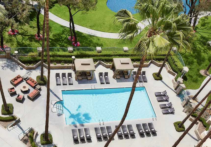 Aerial view of a rectangular outdoor pool surrounded by lounge chairs, cabanas, palm trees, and a nearby fire pit with seating; green lawns and another pool are visible in the background—perfect for relaxing during FIFA World Cup 2026 Los Angeles.