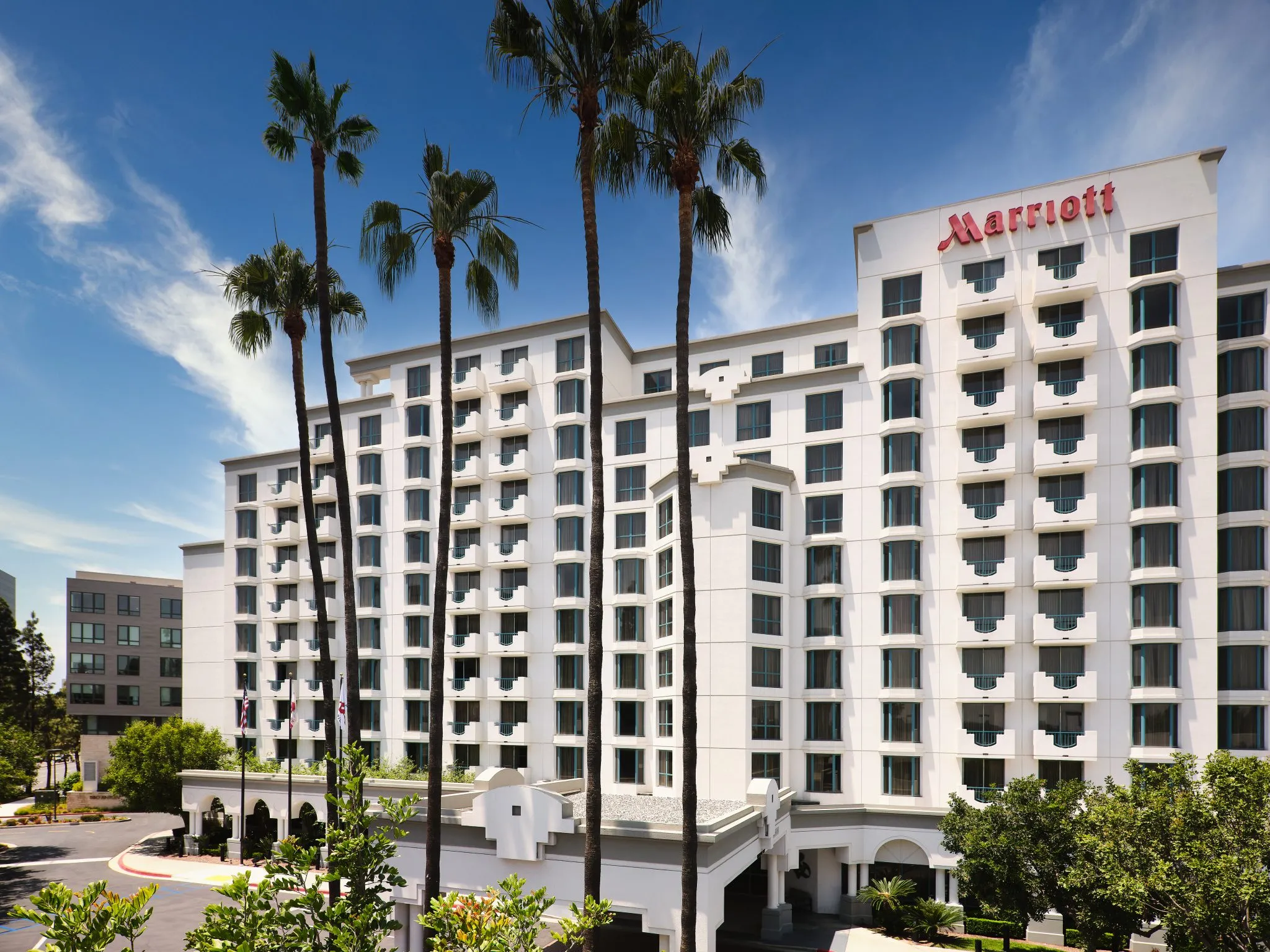 A large white Marriott hotel building with multiple balconies, tall palm trees in front, and a clear blue sky above. The hotel entrance is visible at the bottom center of the image.