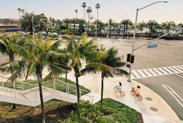 Two people ride bicycles along a palm tree-lined sidewalk near a wide, empty intersection on a sunny day. There are crosswalks, traffic lights, and a street sign reading Coast Hwy.