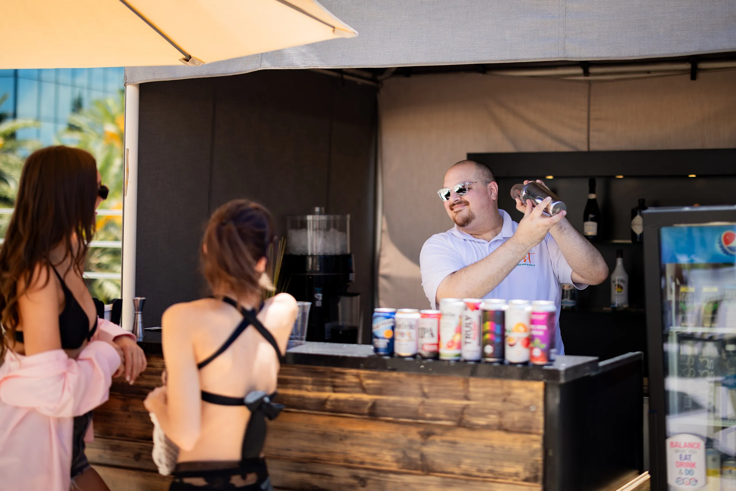 A bartender wearing sunglasses mixes a drink at an outdoor bar while two women in swimsuits stand at the counter. Canned drinks and a refrigerator are visible beside him, with palm trees reflected in the background.