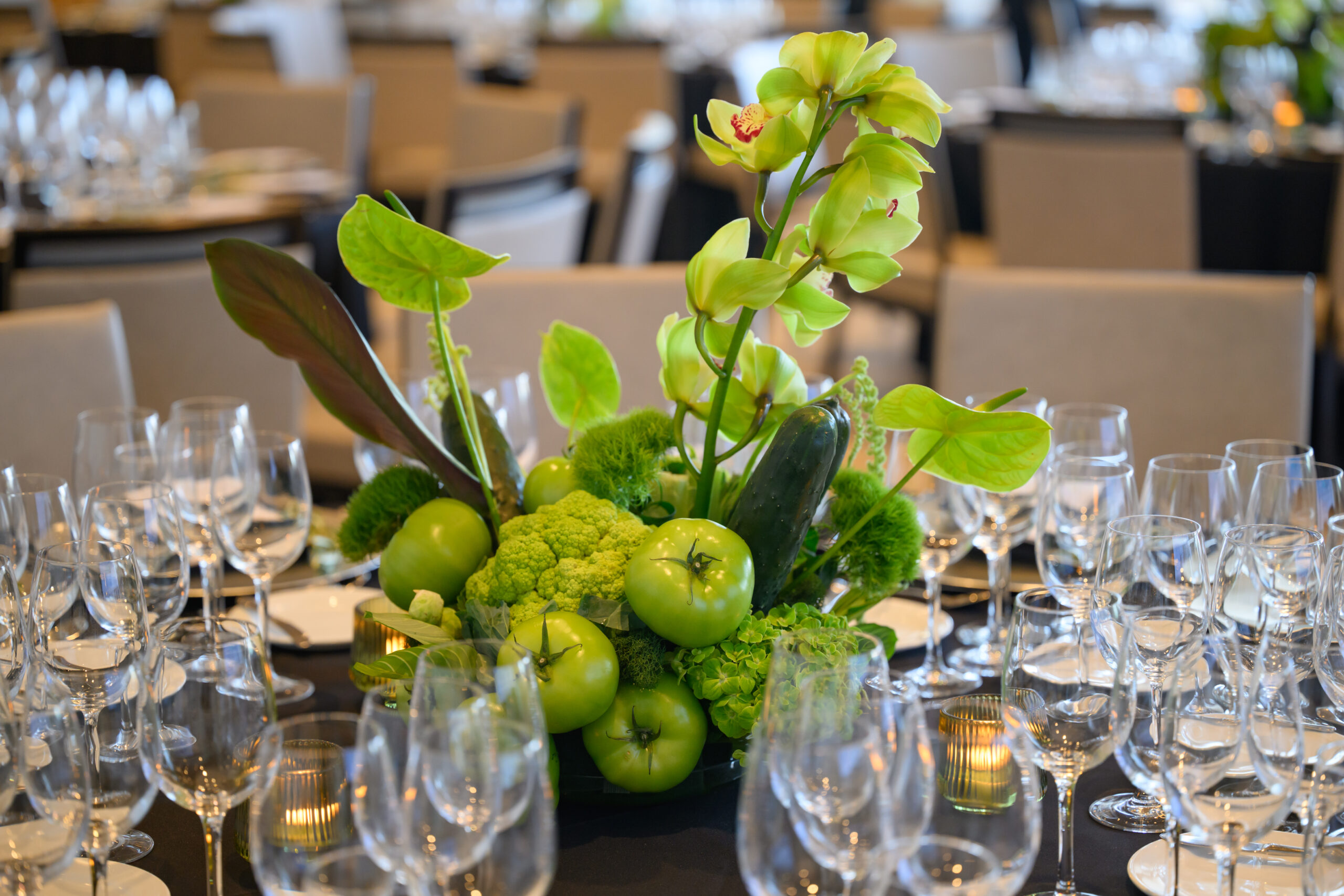 A table centerpiece with green apples, broccoli, cucumbers, and flowers surrounded by empty wine glasses and neatly arranged place settings at a formal dining event.