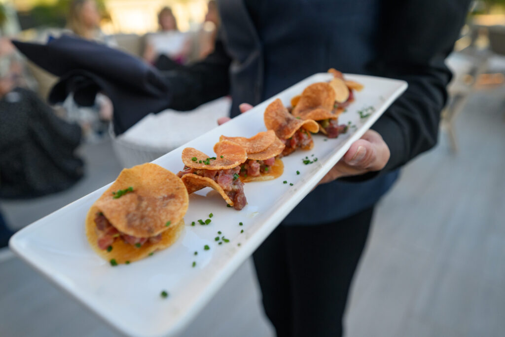 A person in dark clothing holds a rectangular white plate with several small appetizers made of crispy potato chips, garnished with chopped chives.
