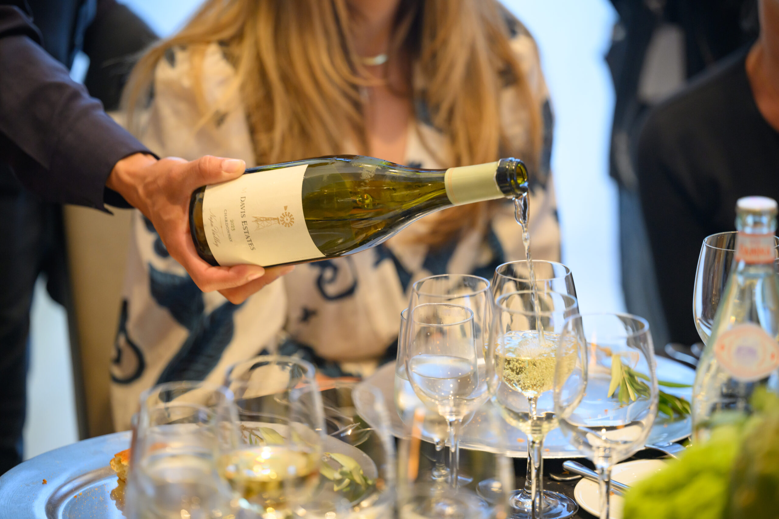 A person pours white wine into a glass at a formal dining table set with plates, glasses, water, and green garnish, while another person sits in the background.