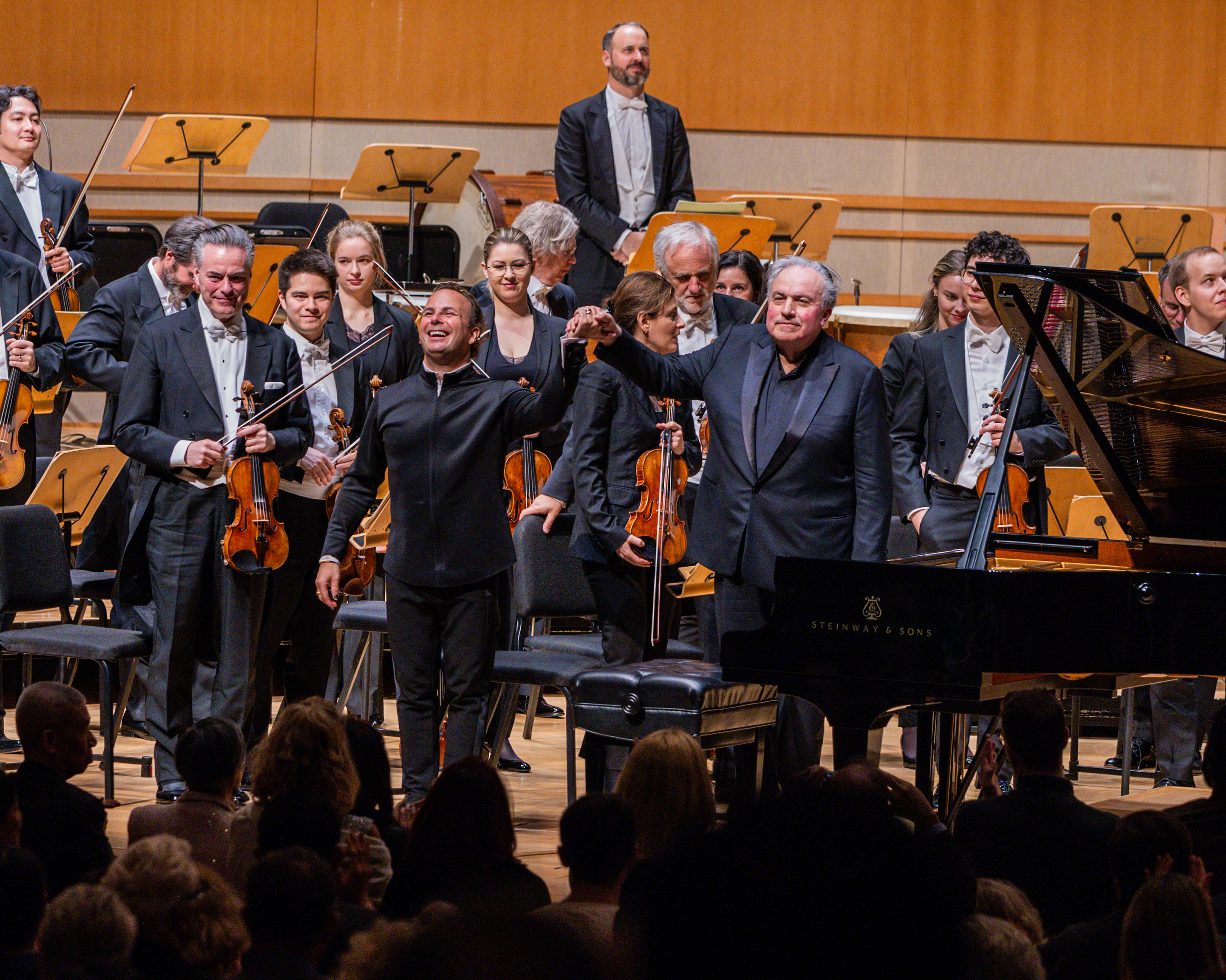 A conductor and a pianist stand in front of a grand piano on stage at Segerstrom Performing Arts Center Costa Mesa, holding hands as they acknowledge the audience. Orchestra members stand behind them with their instruments, smiling and clapping.