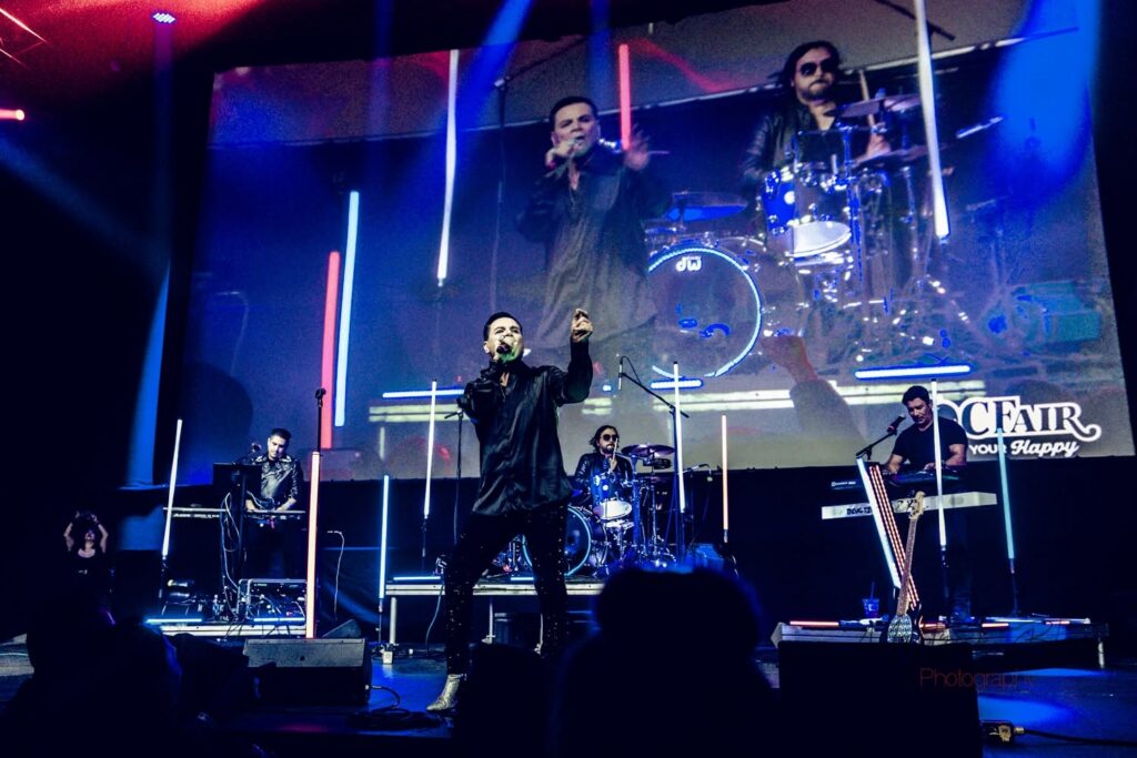 A band performs on stage with bright blue and pink lights, while a large screen behind them shows a close-up of the lead singer. Four musicians are playing keyboards, drums, and guitar. The stage is marked with neon accents.
