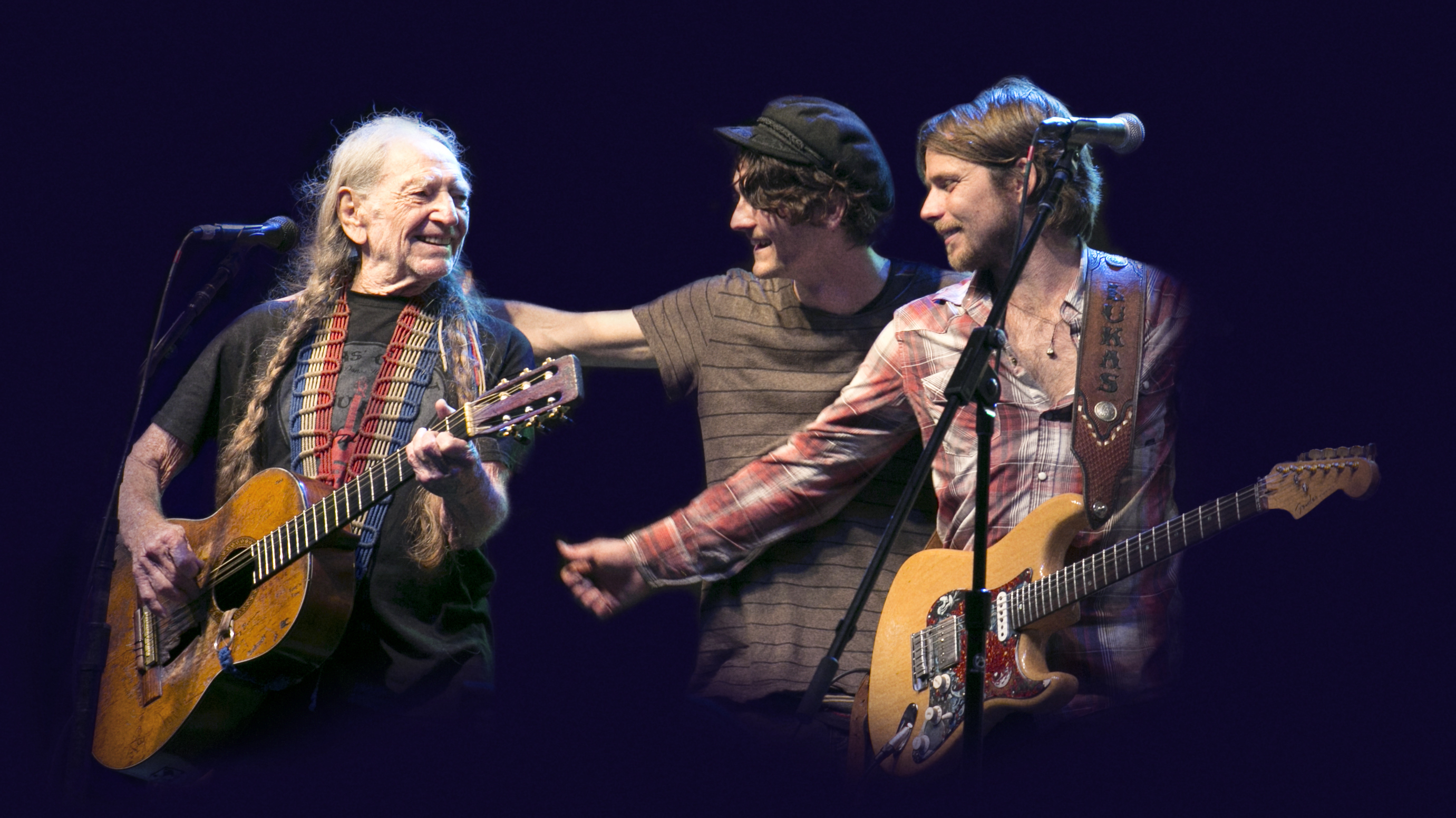 Three musicians perform on stage. One older man plays an acoustic guitar and smiles, while two younger men, one with an electric guitar, interact with each other and the older man. All are under stage lighting with a dark background.