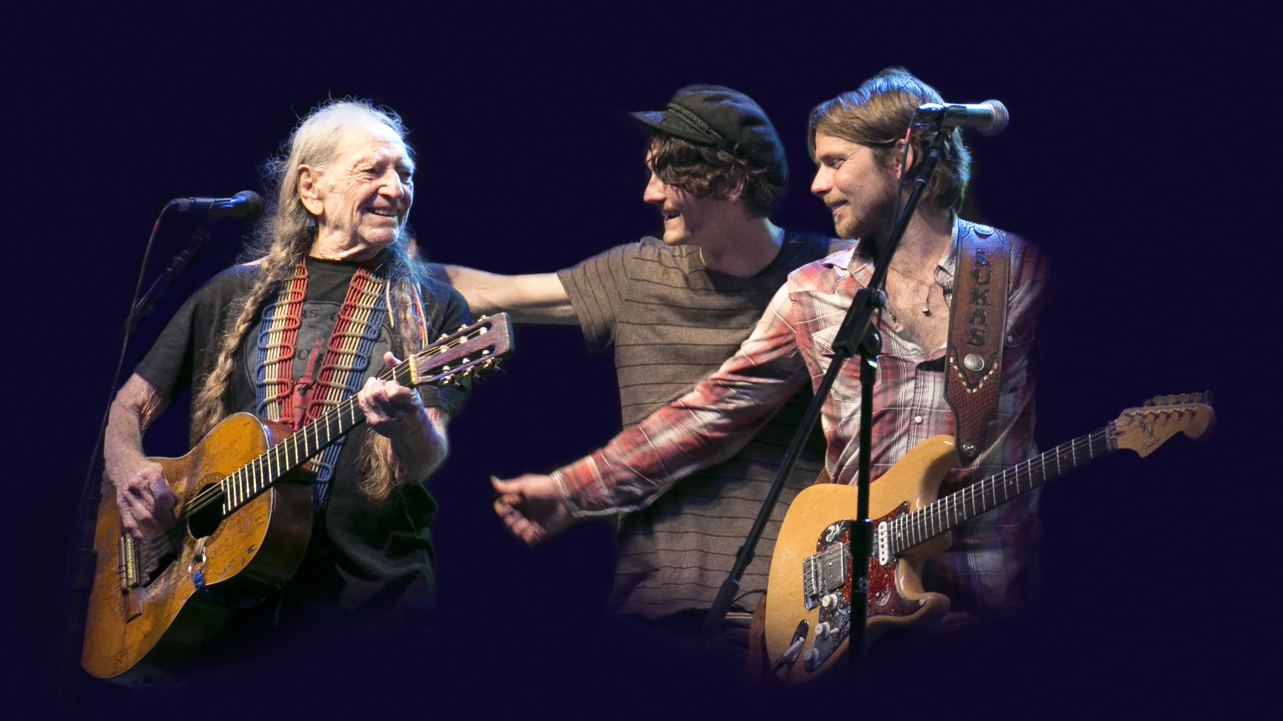 Three musicians perform on stage. One older man plays an acoustic guitar and smiles, while two younger men, one with an electric guitar, interact with each other and the older man. All are under stage lighting with a dark background.