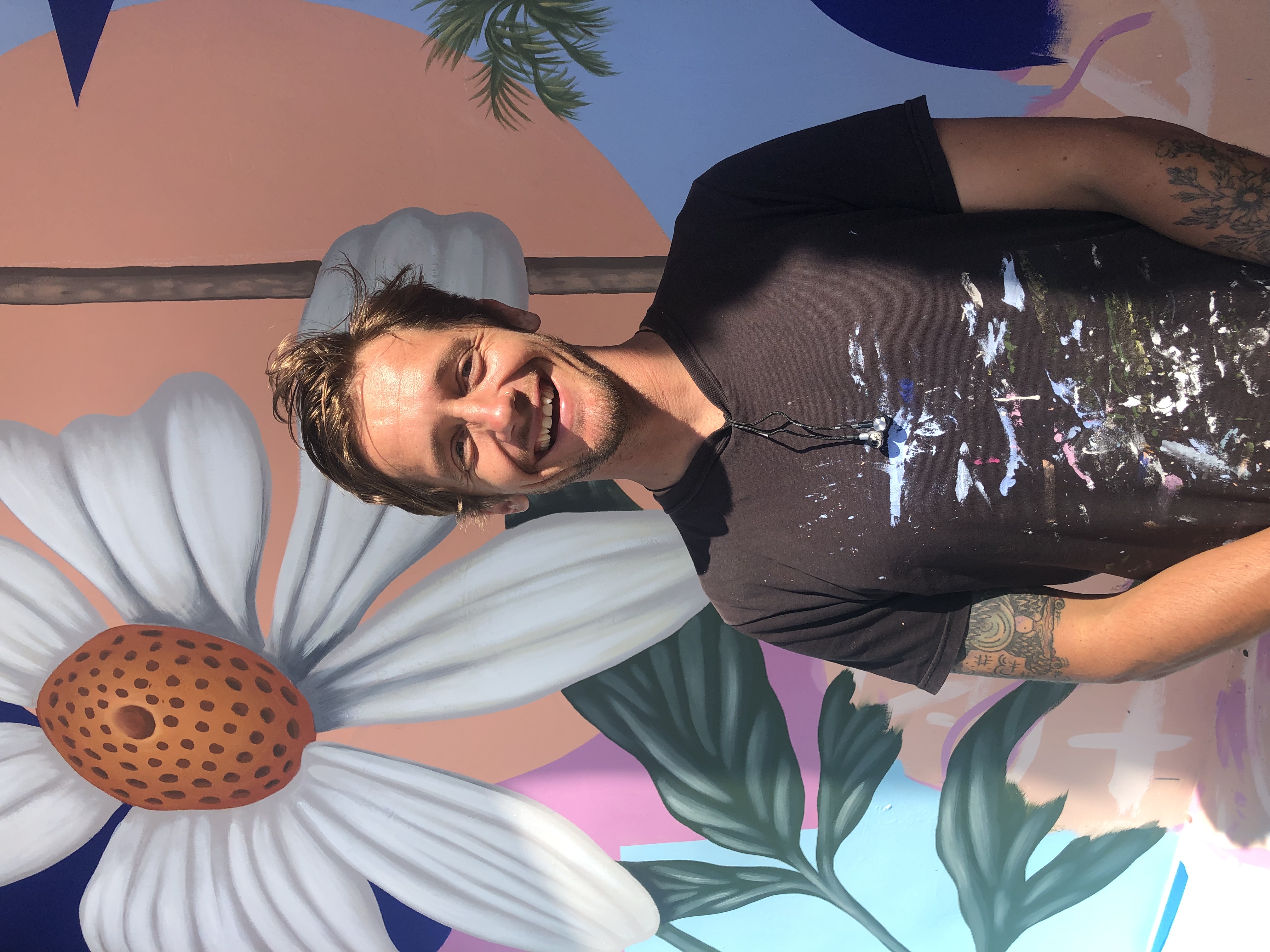 A smiling man with light brown hair stands in front of a colorful mural featuring a large white flower and green leaves. He wears a black t-shirt with paint stains and has tattoos on his left arm.