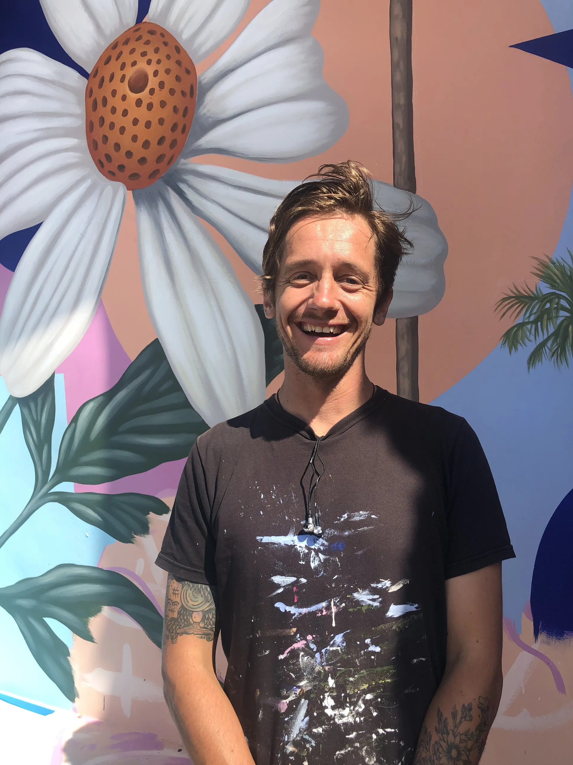 A smiling man with light brown hair stands in front of a colorful mural featuring a large white flower and green leaves. He wears a black t-shirt with paint stains and has tattoos on his left arm.