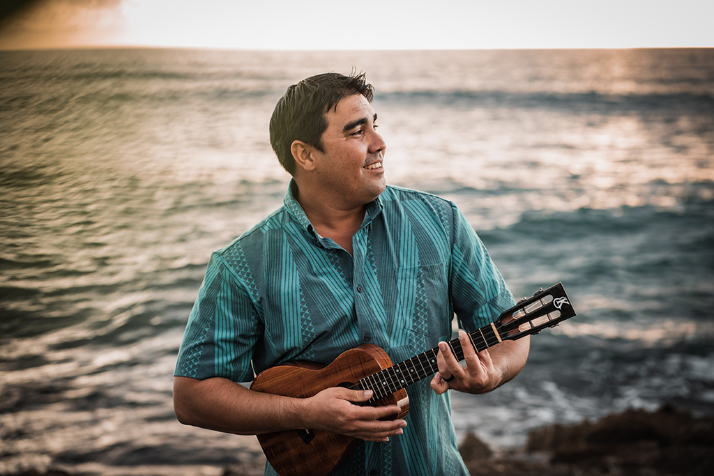 A man in a blue patterned shirt smiles while playing a ukulele on a rocky shore with the ocean and sunset in the background.