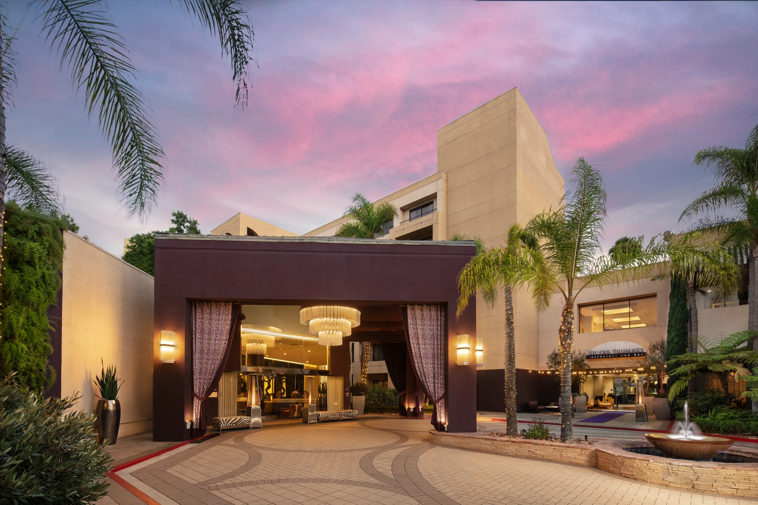 Hotel entrance at sunset with palm trees, modern architecture, a covered driveway, and chandeliers hanging above the glass doors. The sky is pink and purple, creating a welcoming and elegant atmosphere.