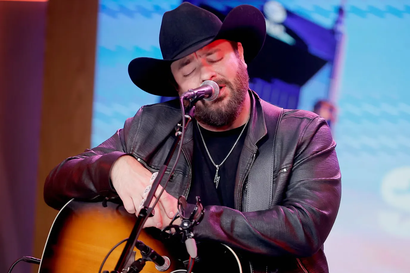 A man wearing a black cowboy hat and leather jacket plays an acoustic guitar and sings into a microphone on stage, with his eyes closed.