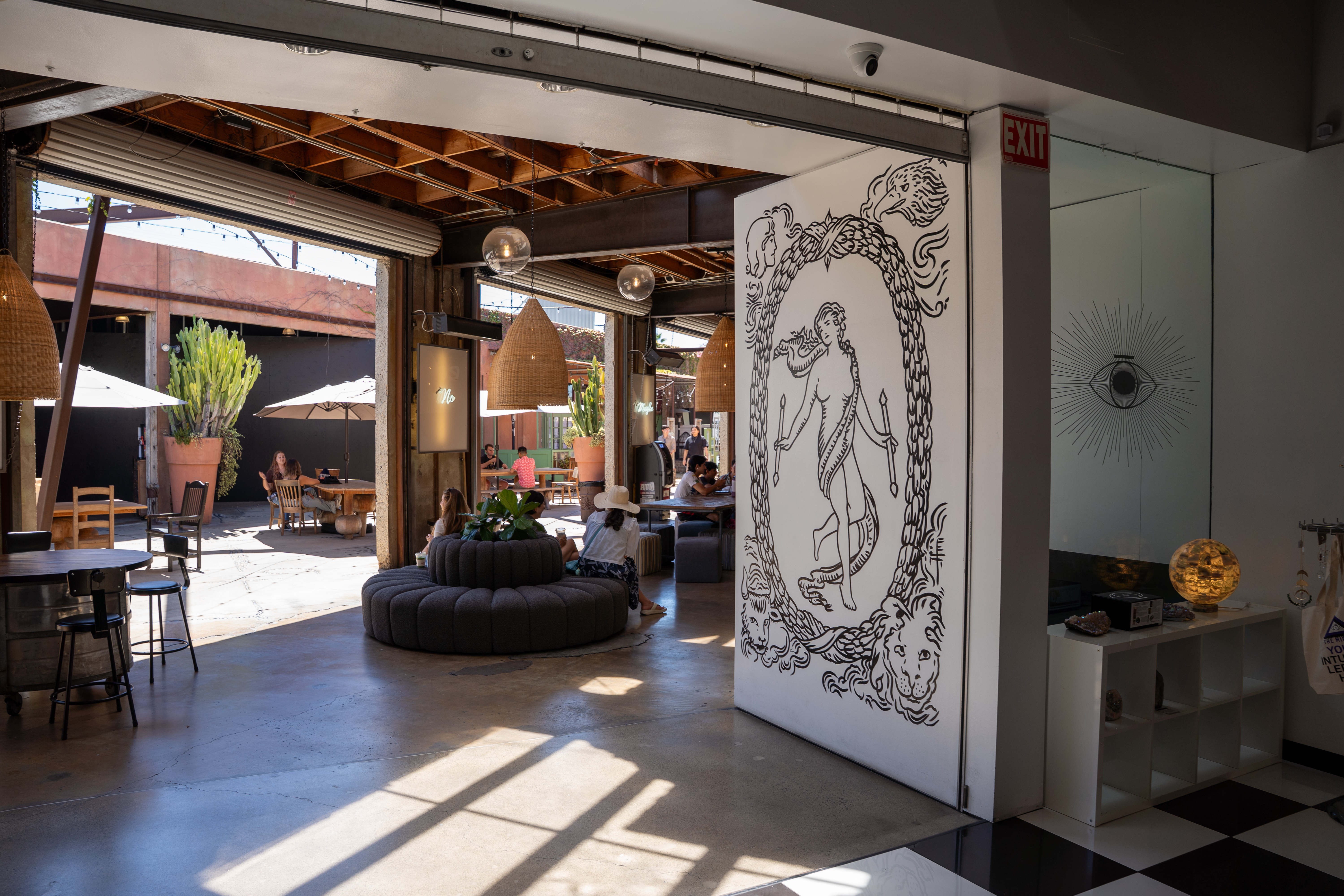 A modern indoor-outdoor café space in a shopping mall Costa Mesa, filled with natural light, wooden furniture, lush plants, and a white wall with a striking black mural. People are sitting and relaxing both inside and outside.