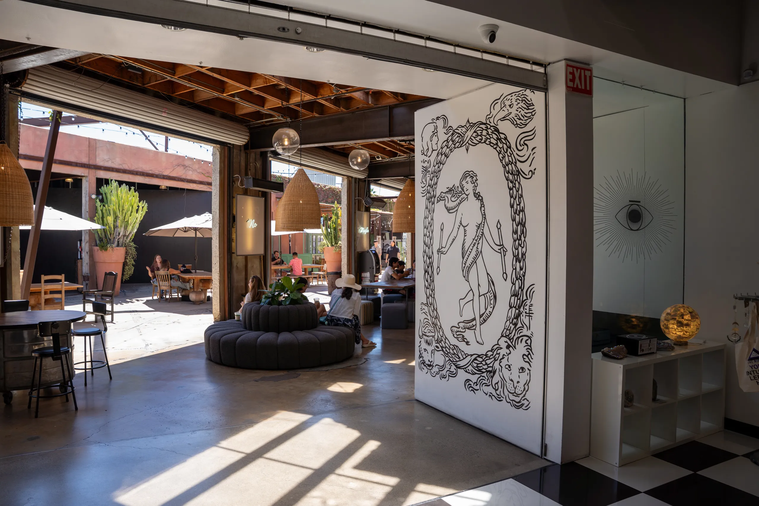A modern indoor-outdoor café space in a shopping mall Costa Mesa, filled with natural light, wooden furniture, lush plants, and a white wall with a striking black mural. People are sitting and relaxing both inside and outside.
