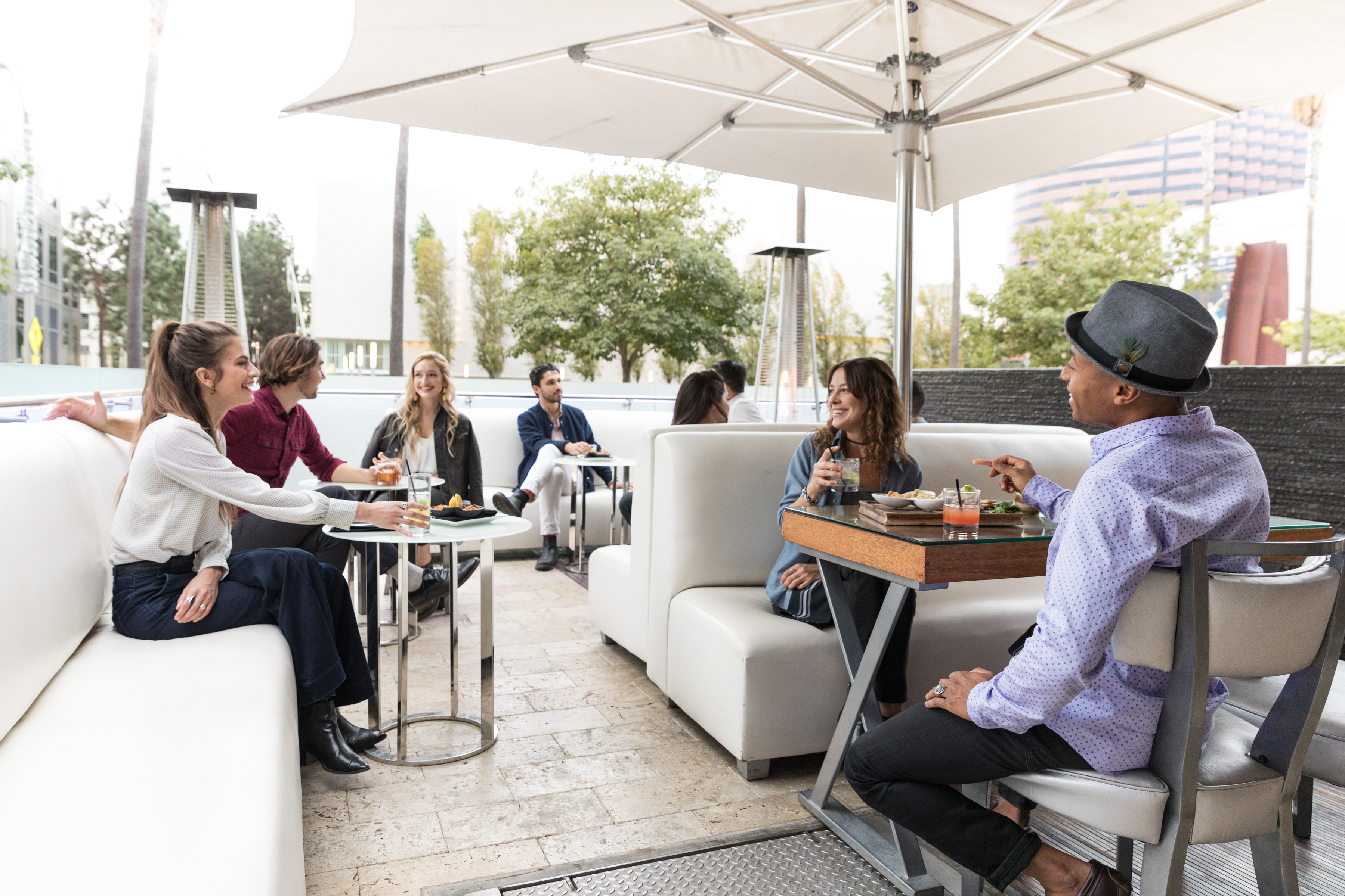 A group of people sit at outdoor restaurant tables, talking and eating under a large white umbrella. Some have drinks, and everyone appears relaxed and engaged in conversation. Trees and buildings are visible in the background.