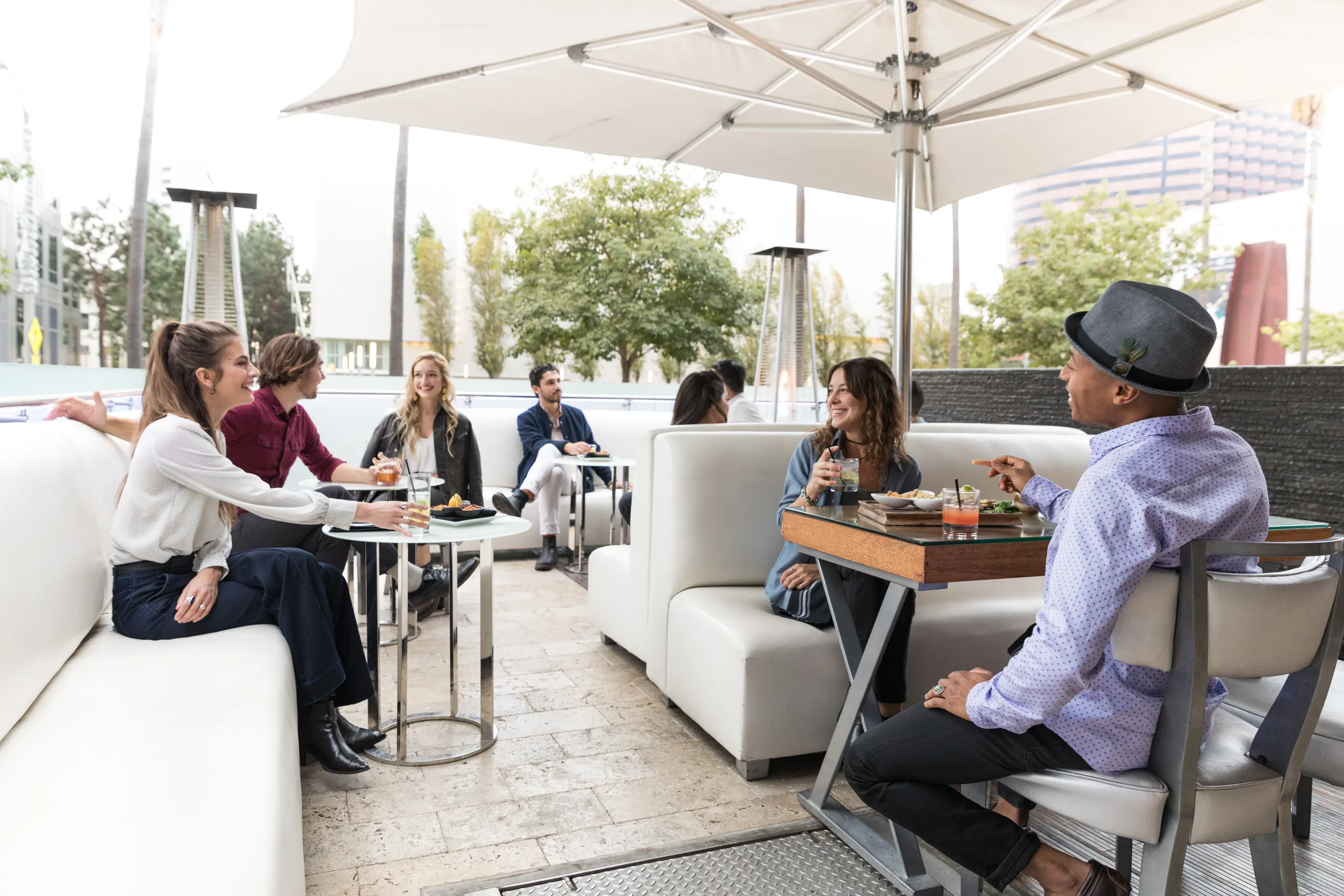A group of people sit at outdoor restaurant tables, talking and eating under a large white umbrella. Some have drinks, and everyone appears relaxed and engaged in conversation. Trees and buildings are visible in the background.