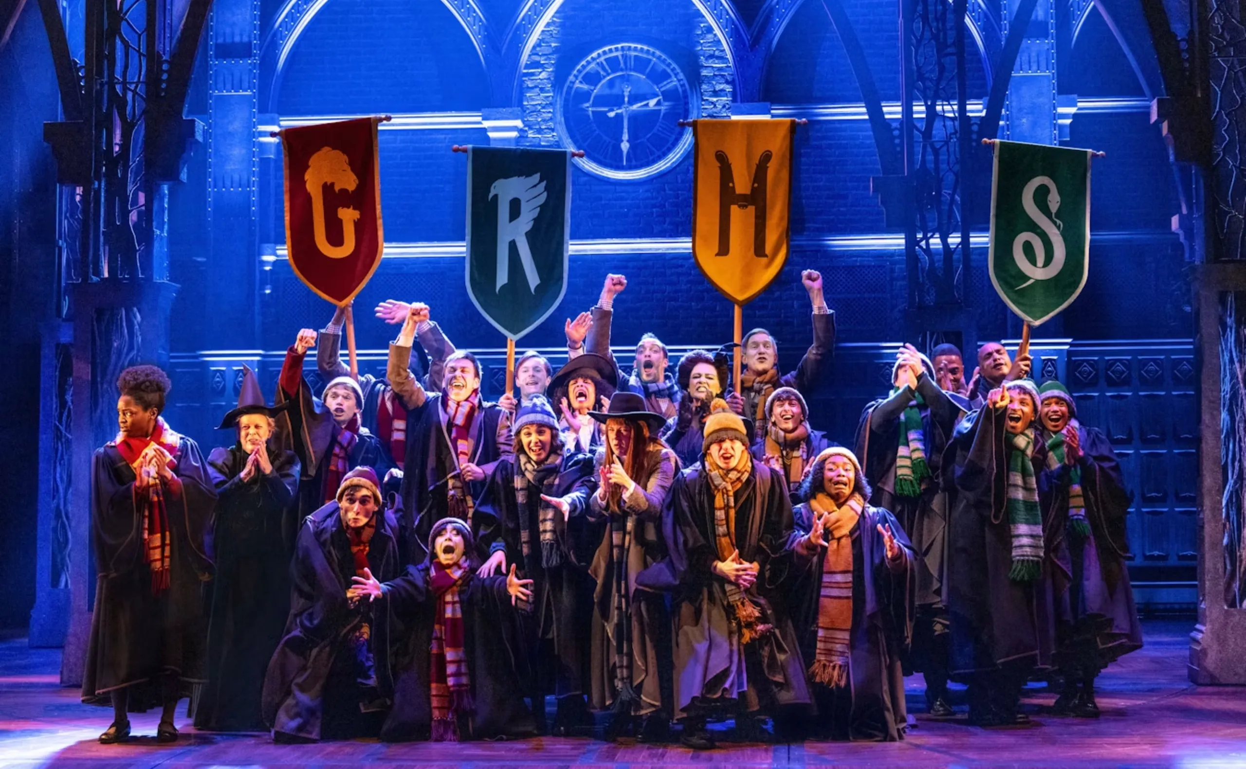 A group of people dressed as Hogwarts students and professors on stage, cheering and holding colorful house banners for Gryffindor, Ravenclaw, Hufflepuff, and Slytherin, with a magical, blue-lit set behind them.