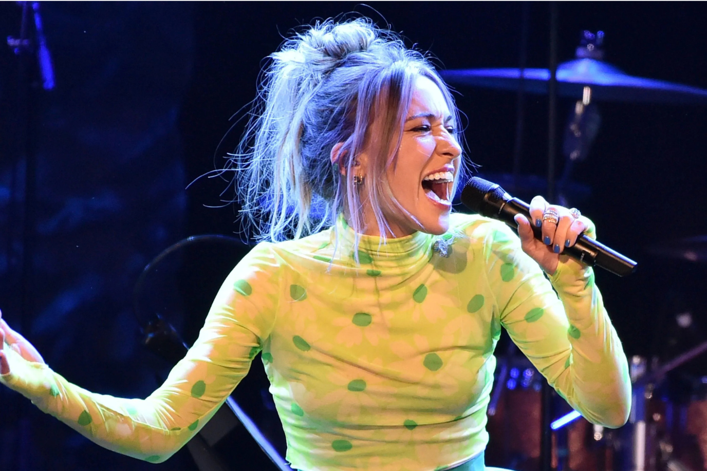 A woman with a high messy bun and wearing a bright green, long-sleeve, polka dot top passionately sings into a microphone on stage, with stage lights and drum equipment visible in the background.
