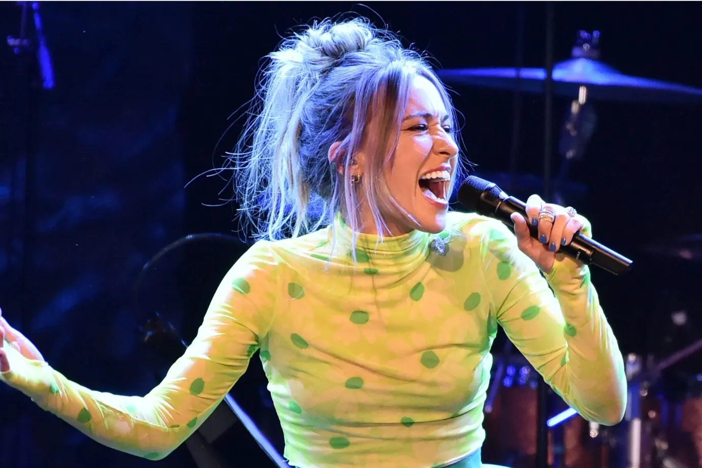 A woman with a high messy bun and wearing a bright green, long-sleeve, polka dot top passionately sings into a microphone on stage, with stage lights and drum equipment visible in the background.