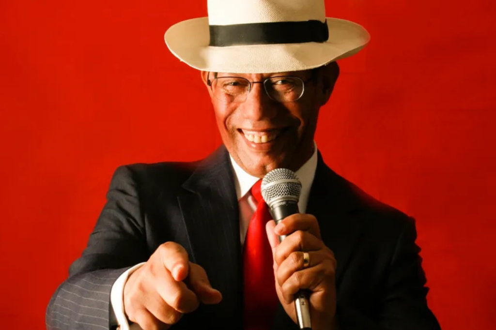 A man in a suit, red tie, and white fedora smiles while holding a microphone and pointing toward the camera, standing in front of a bright red background.