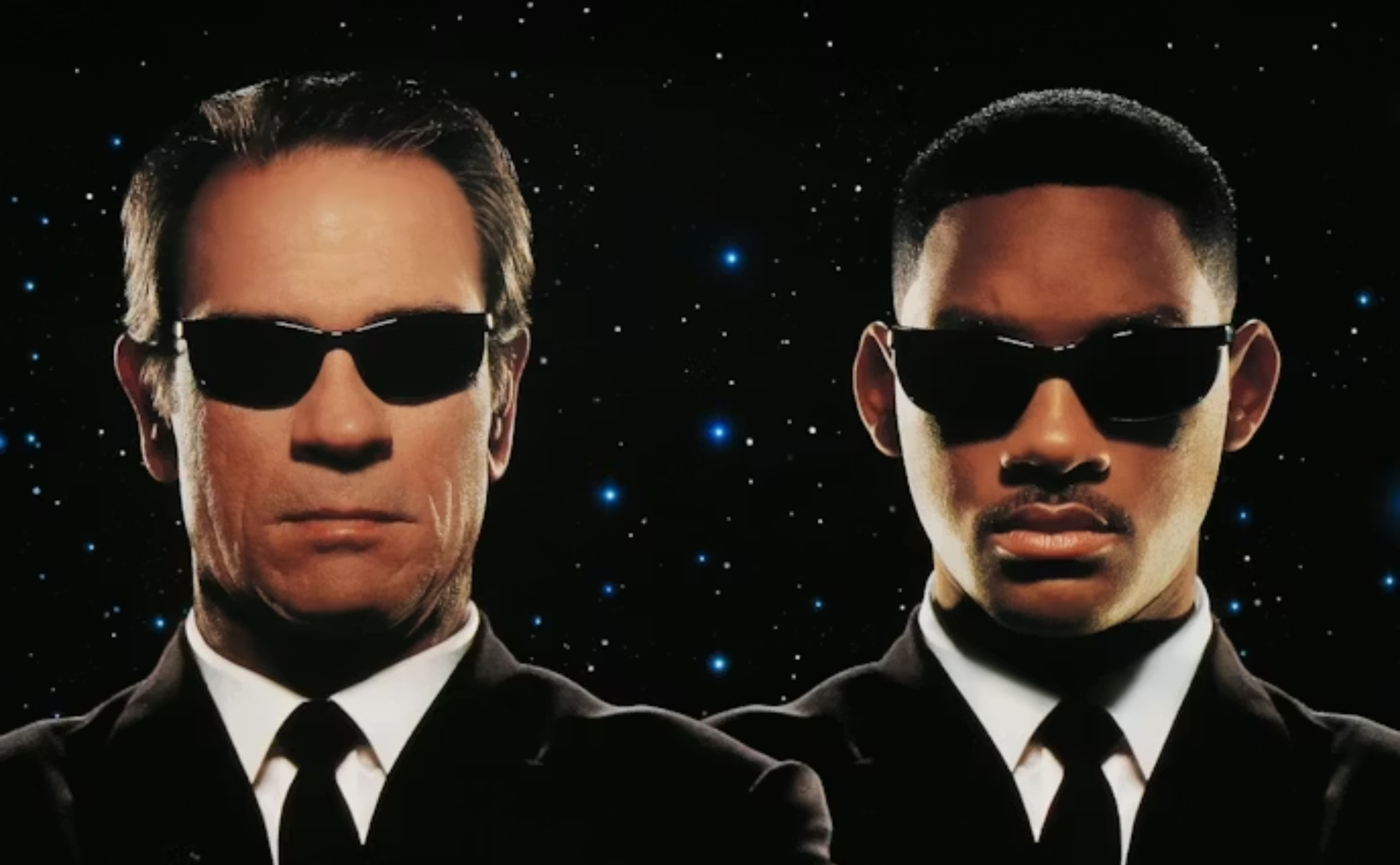 Two men wearing black suits, white shirts, black ties, and dark sunglasses stand side by side against a black background with small blue stars. They both have serious expressions.