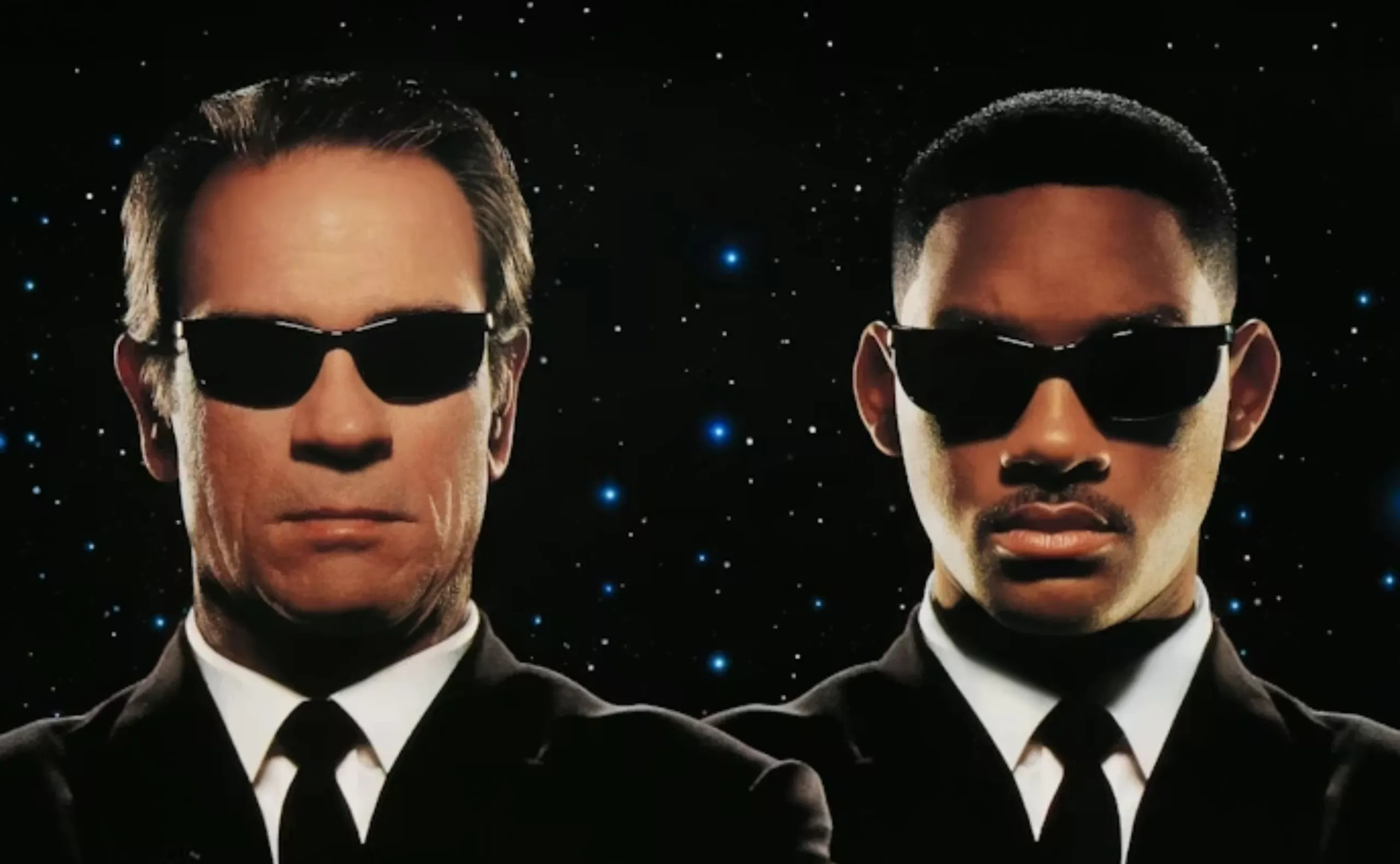 Two men wearing black suits, white shirts, black ties, and dark sunglasses stand side by side against a black background with small blue stars. They both have serious expressions.