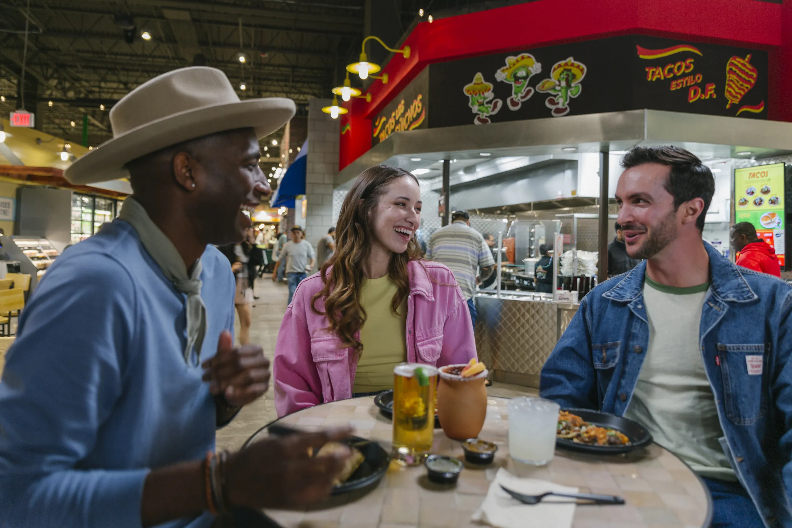 Three people sit at a table in a casual restaurant, smiling and talking while enjoying food and drinks. A vibrant taco stand is visible in the background, adding a lively atmosphere.