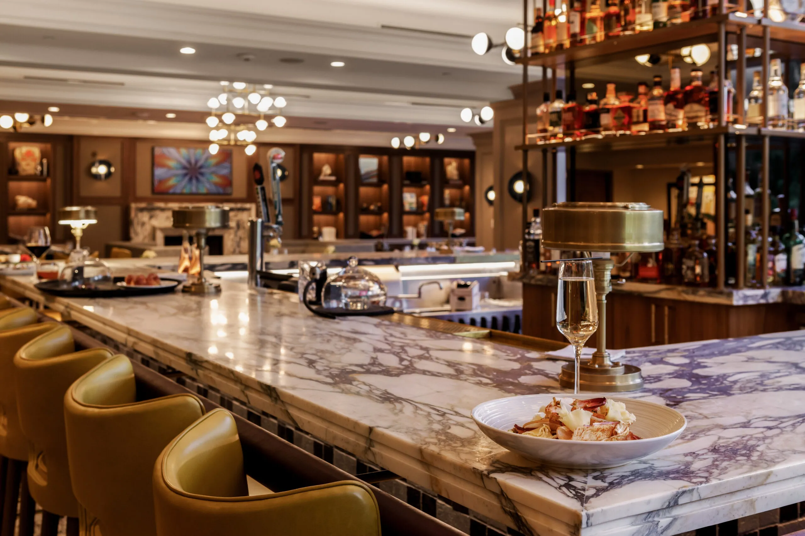 A marble bar with yellow stools, a plate of food, and a glass of wine in the foreground. Shelves of liquor bottles and warm lighting decorate the background, creating a cozy, upscale atmosphere.