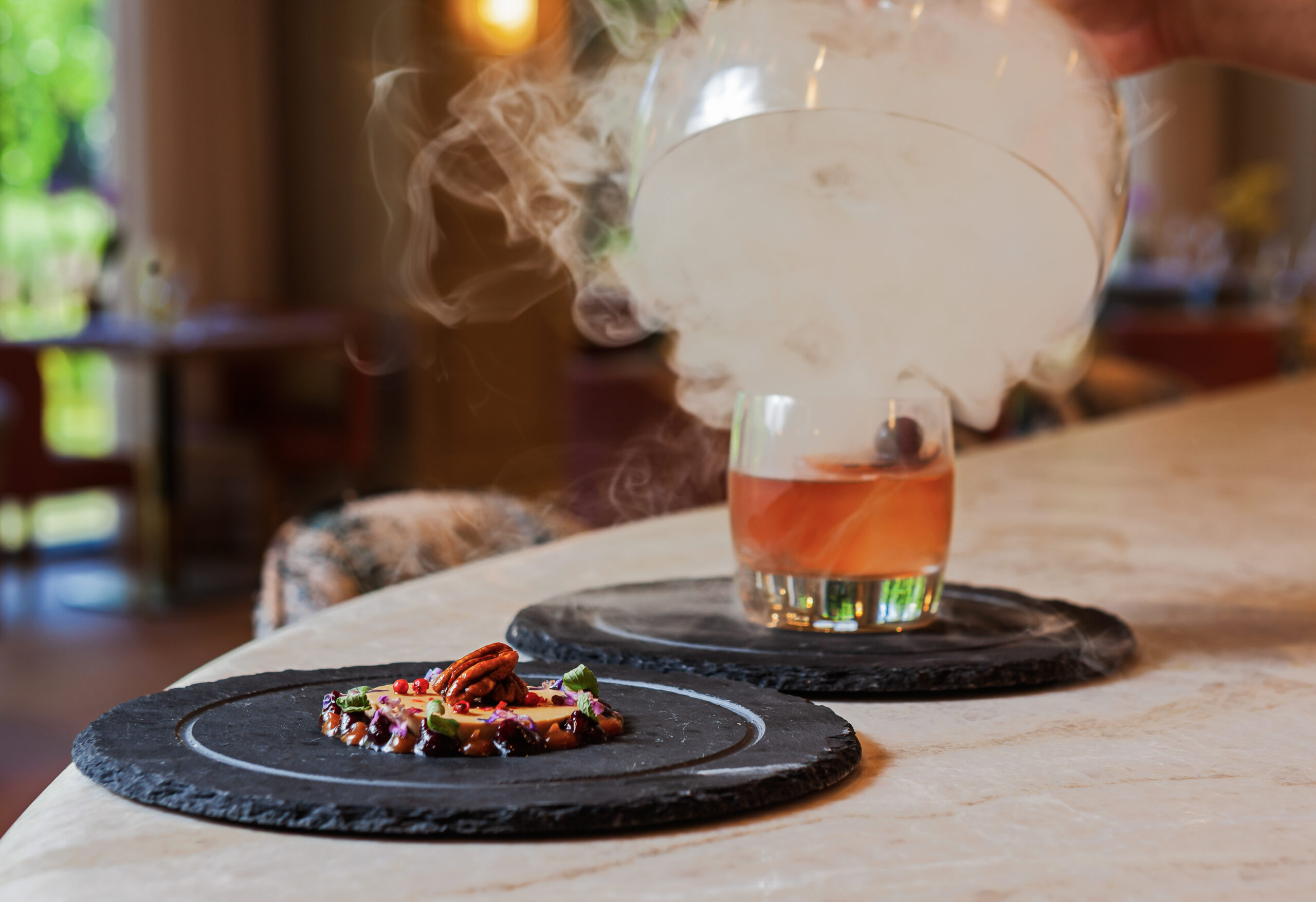 A smoked cocktail is revealed as a glass cover is lifted, with a gourmet appetizer garnished with pecans on a black slate plate nearby, set on a marble countertop.
