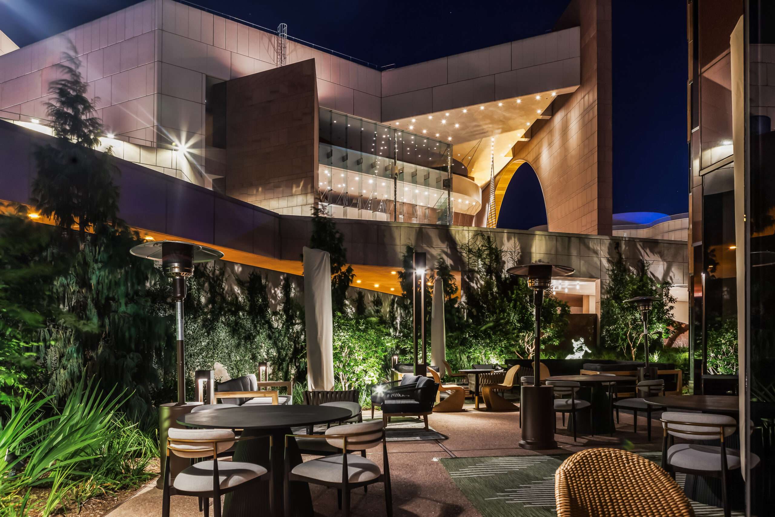 Modern outdoor patio at night with tables, chairs, greenery, and overhead heaters, set against a backdrop of a contemporary building with illuminated balconies and geometric architecture.
