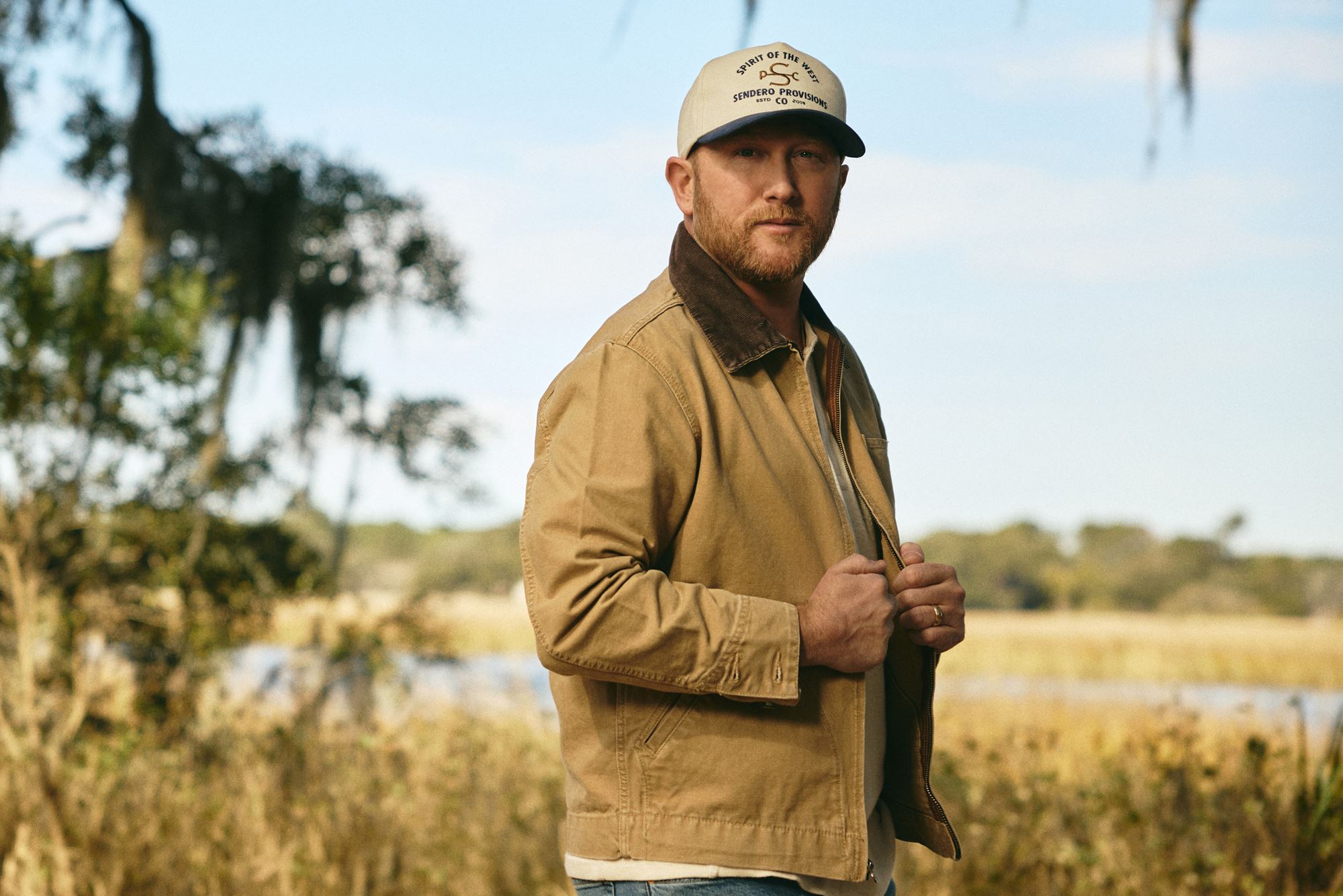 A man stands outdoors in a field near water, wearing a tan jacket and a light-colored baseball cap. Trees and tall grass are visible in the background under a blue sky.